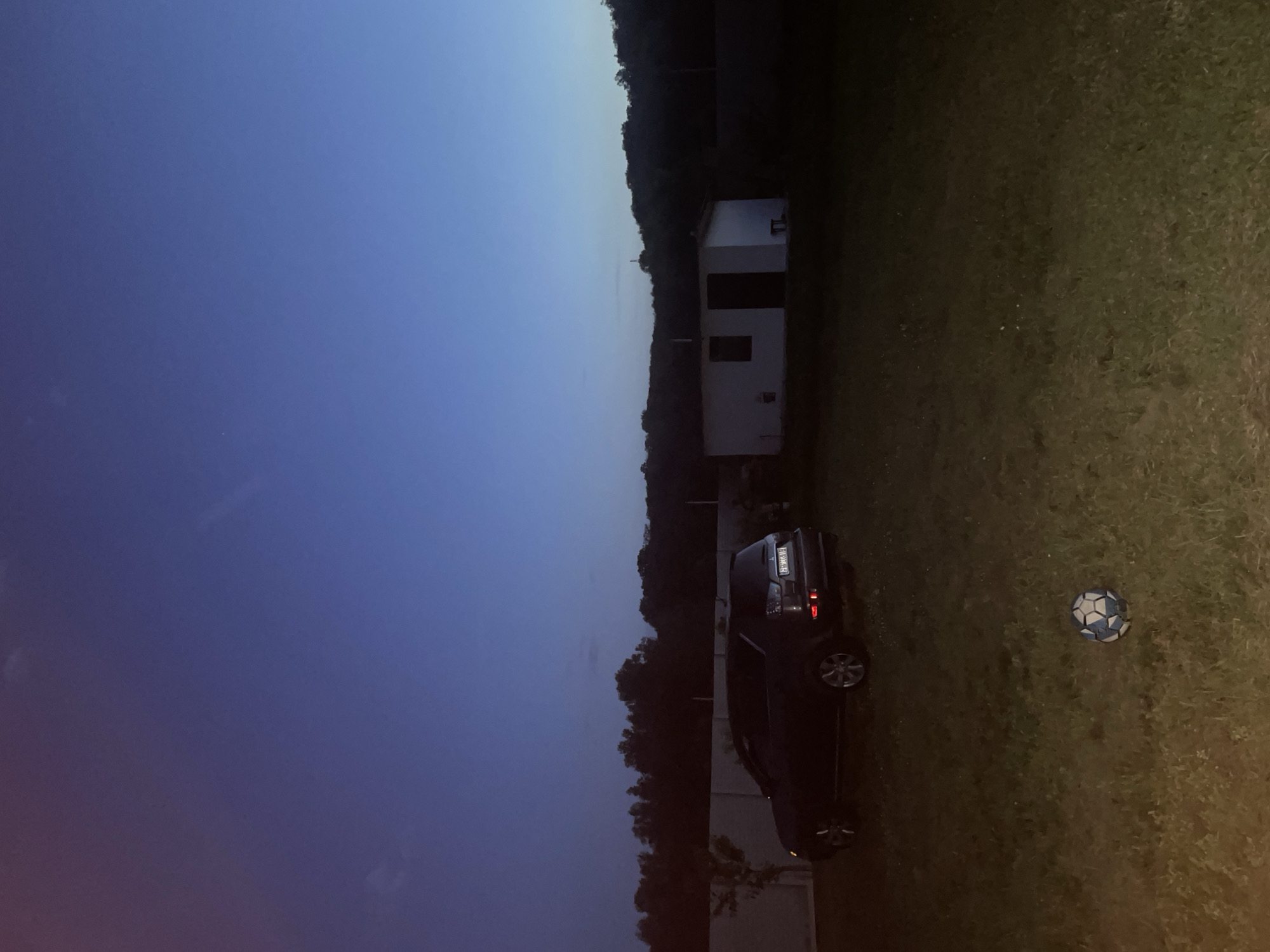 The scene depicts a twilight or early night landscape in Petrivka, Ukraine. The sky, occupying the upper two-thirds of the image, presents a deep indigo blue at the zenith, gradually transitioning to a lighter blue-grey near the horizon, with subtle hints of red-purple haze on the far left. The foreground consists of a sparsely mown grassy area, illuminated by an unseen light source, casting a yellowish-green hue on the ground. A blue and white patterned soccer ball rests on the grass in the lower center.
In the midground, a dark-colored SUV, consistent with a Mitsubishi Outlander, is parked facing right on the grassy terrain. Its rear features illuminated taillights and a visible Ukrainian license plate, reading "BH 0051 IB", indicating the Odesa region. Behind the SUV, a light-colored, possibly concrete, perimeter fence stretches horizontally across the scene. Small, dark, sparse vegetation and trees are visible both behind and in front of the fence line on the left.
To the right of the SUV and fence, a small, white, rectangular, single-story utility building or shed is visible, featuring a dark door and a rectangular window. A small, dark object, resembling a bench or stool, sits directly in front of the building's right side. Behind this structure and extending across the right side of the frame is a dense line of dark, silhouetted trees, suggesting a wooded area or forest. The overall atmosphere is devoid of human presence or activity.
