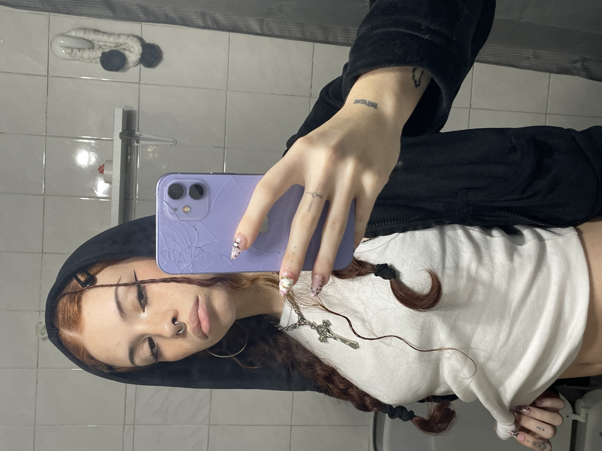 The image shows a young person with red-brown braided hair, wearing a black hoodie and a white top. They have a nose ring and are holding a purple smartphone with a cracked screen. The person is likely taking a selfie in a bathroom, indicated by the tiled wall, mirror, shower fixture, and a towel holder in the background. The person has a cross necklace and several tattoos on their hands and wrist. The exact location in Buenos Aires, Argentina cannot be determined from the given image.