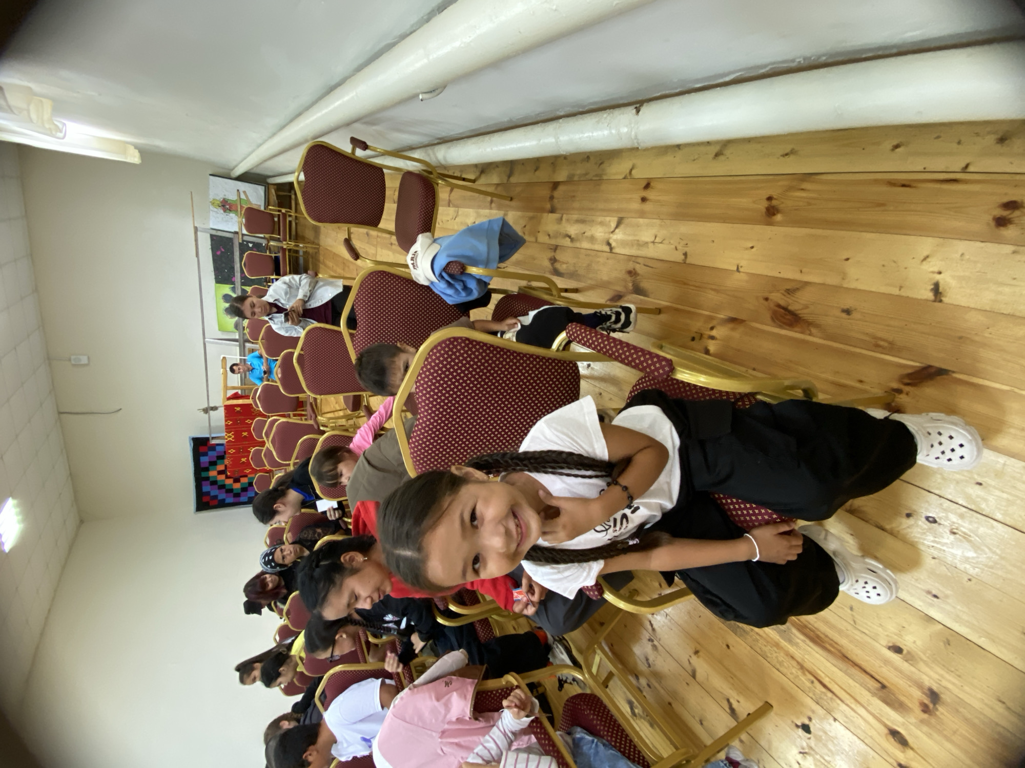 A group of children and adults are seated in a room with rows of chairs, some of which are empty. The room has a wooden floor and white walls. In the foreground, a young girl with braided dark hair and wearing a white t-shirt and black pants smiles at the camera. She is sitting in a red and gold patterned chair. Behind her, other individuals are visible, some looking towards the camera and others looking in different directions. In the background, there are paintings and decorations on the wall, and more chairs are arranged in rows. The overall scene appears to be an indoor gathering or event, possibly in a community hall or school in Maloarslangulovo, Russia.