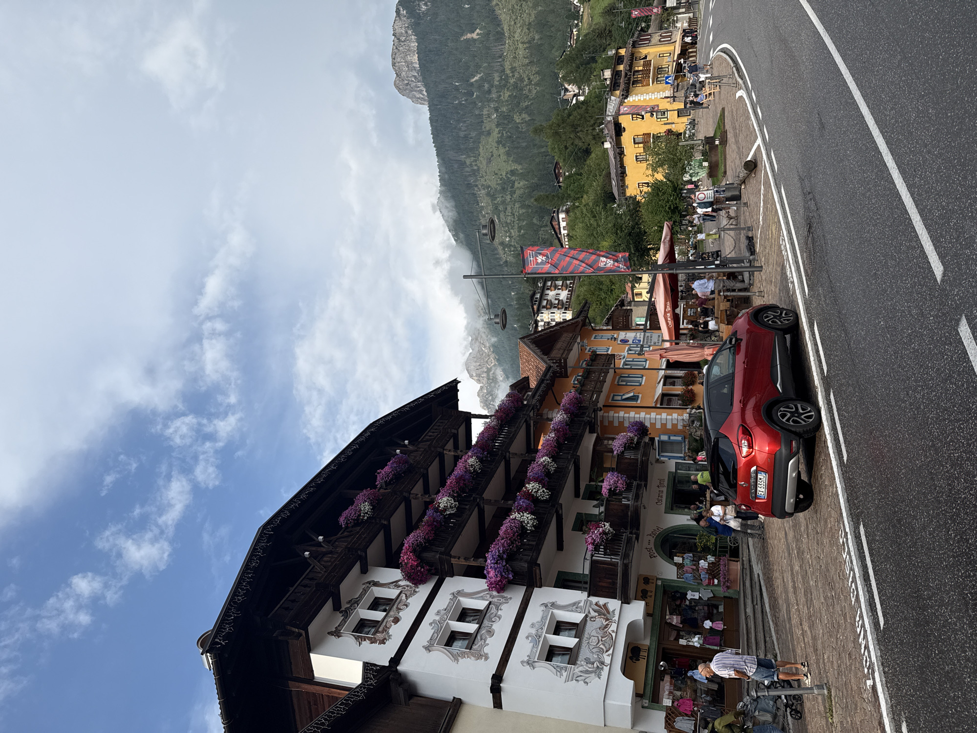 A red Renault Captur is parked on the right side of a paved road in Moena, Italy, with its license plate reading "EX 535MJ". The road has white lane markings. In the background, a white building with dark wood trim and balconies overflowing with purple and pink flowers stands prominently. Below the balconies, storefronts with awnings are visible, including one labeled "Post Hotel Ostaria Tirol". To the right of this building are structures painted in yellow and orange hues, also featuring wooden balconies. People are scattered along the sidewalk, some walking, others standing near the buildings. A mountainous landscape with dense green trees and rocky peaks shrouded in mist rises behind the town. The sky is partly cloudy with a mix of blue and white.