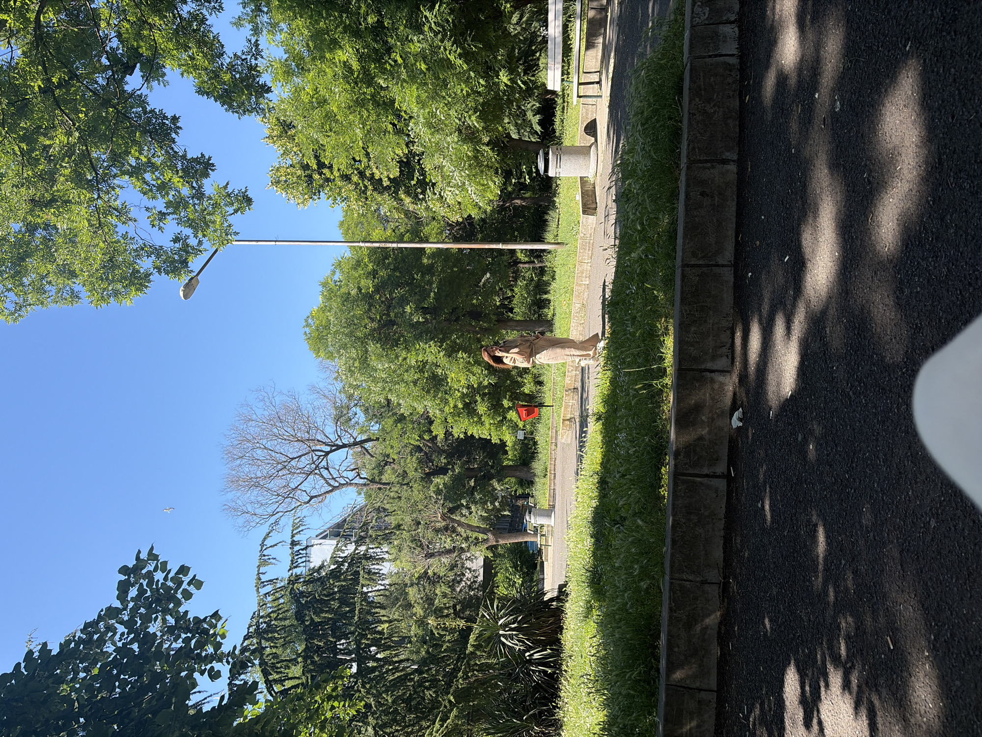 The image depicts a sunny day in a park in Burgas, Bulgaria. A woman wearing a beige jumpsuit is standing and talking on her phone in the middle of a paved path. The path is bordered by a strip of grass and then a low concrete wall. Beyond the path, there are trees, a lamppost, a trash can, and a bench. In the distance, a building is visible. The sky is clear and blue, and the sun casts shadows on the ground. The overall impression is of a peaceful park scene on a warm day.