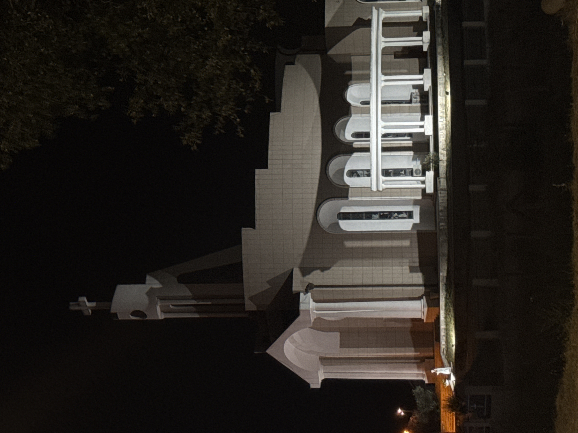 The image depicts a religious building, possibly a church, at night in Donji Štoj - Shtoji i Poshtëm, Montenegro. The structure has a prominent cross atop its steeple. The building is illuminated, casting shadows. A portico with columns is visible in front of the main structure, featuring arched windows. In front of the building, a small, solitary figure, likely a statue, stands on a raised platform. The surrounding environment is dark, with some foliage visible on the right side of the image. The foreground is obscured by darkness, with hints of grass and what appears to be a low barrier or fence.