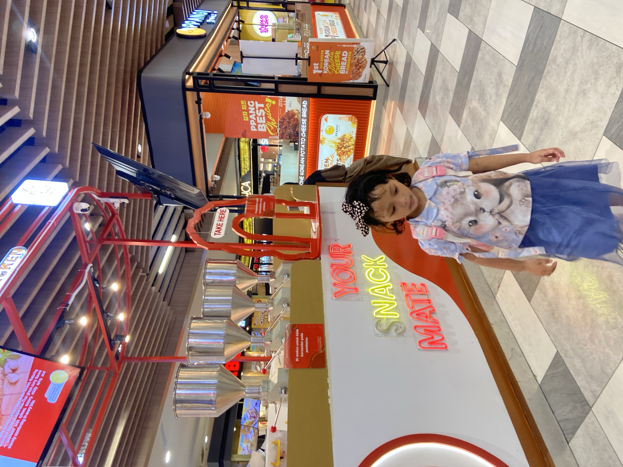 The image depicts an indoor scene within what appears to be a modern shopping mall or food court in Surabaya, Indonesia. A young girl, likely of elementary school age, is the main human subject, positioned on the right side of the frame. She has dark hair with a polka-dotted headband and is wearing a light blue shirt adorned with a cat graphic and a dark blue, possibly tulle, skirt. Her expression is subtle, looking towards the left.
In the foreground, a brightly lit snack dispensing station, possibly for bulk or self-serve snacks, features several metallic, funnel-shaped containers with levers. A prominent neon sign on the station reads "YOUR SNACK MATE" in red and yellow letters. Another sign advises "TAKE HERE." A red instruction card, partially visible, states in Indonesian, "Di mohon untuk tidak bersandar pada pembatas" (Please do not lean on the barrier).
The background reveals several food kiosks. One prominent stall, identified by signs such as "PPANGI" and "ONE KOREAN POTATO CHEESE BREAD," proudly declares itself the "1st KOREAN Potato CHEESE BREAD IN SURABAYA." Other visible text includes "PPANG BEST Choice" and "shake shake." Marketing materials display tempting images of food, with descriptions like "감자 치즈" (Potato Cheese in Korean) and details about "Japanese-Thai green curry flavors." The environment is well-lit, suggesting daytime. The ceiling above features a contemporary design with wooden slats, while the floor is tiled with a contrasting geometric pattern of light and dark grey squares.