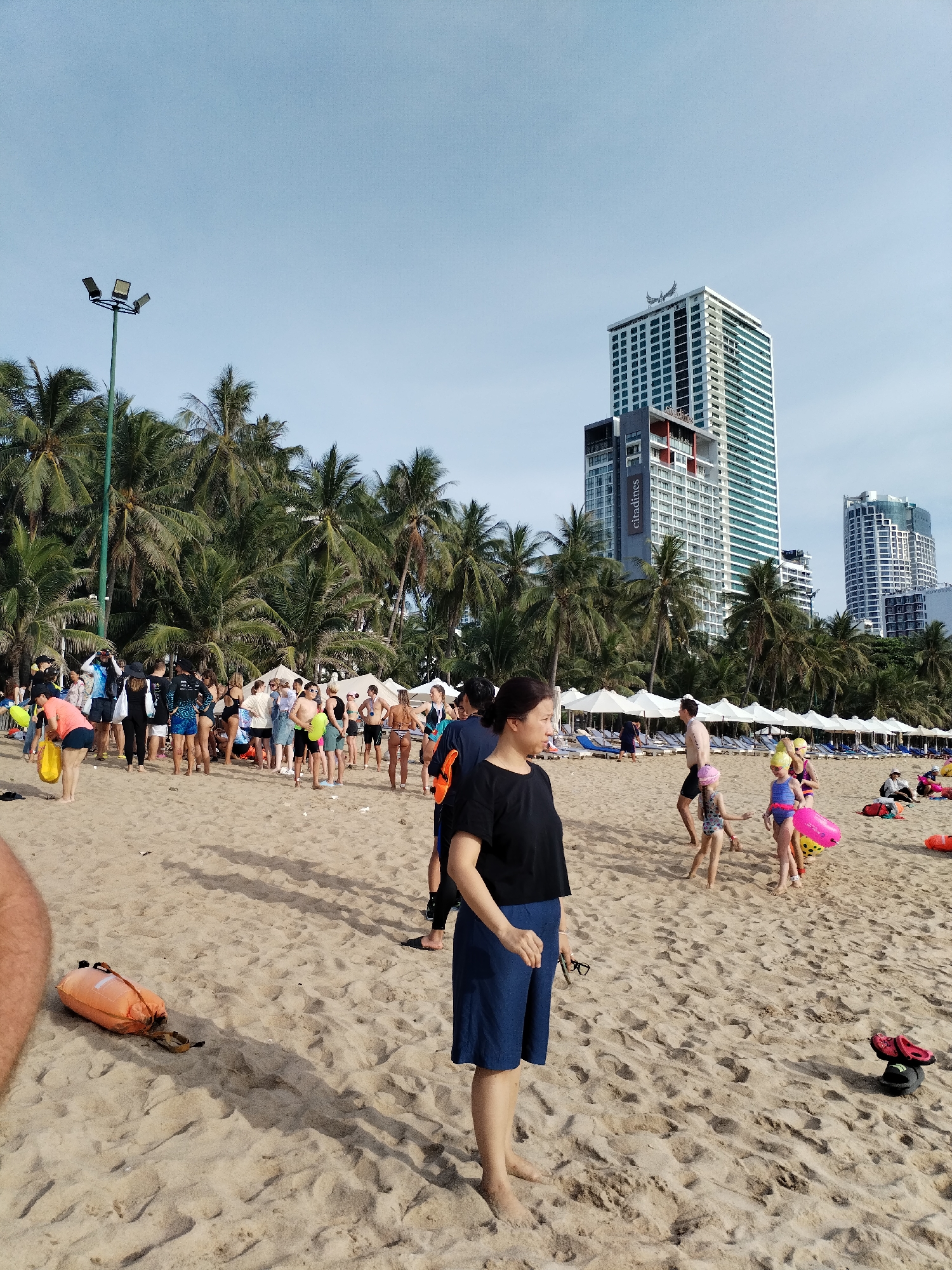 This image depicts a crowded beach in Nha Trang, Vietnam. In the foreground, a woman wearing a black t-shirt and blue shorts stands facing right, her feet bare on the sand. To her left, an orange inflatable buoy lies on the beach. In the mid-ground, a group of people, including children in swimsuits and some adults, are gathered. Some children are playing with a pink inflatable toy. Further back, a large number of people are standing and milling about on the sand, with palm trees and white beach umbrellas in the background. Behind the beach, several tall buildings, characteristic of Nha Trang's skyline, are visible, including a prominent white and green high-rise with the "citadines" logo. The sky is clear and blue. The overall scene suggests a busy day at a popular beach destination.
