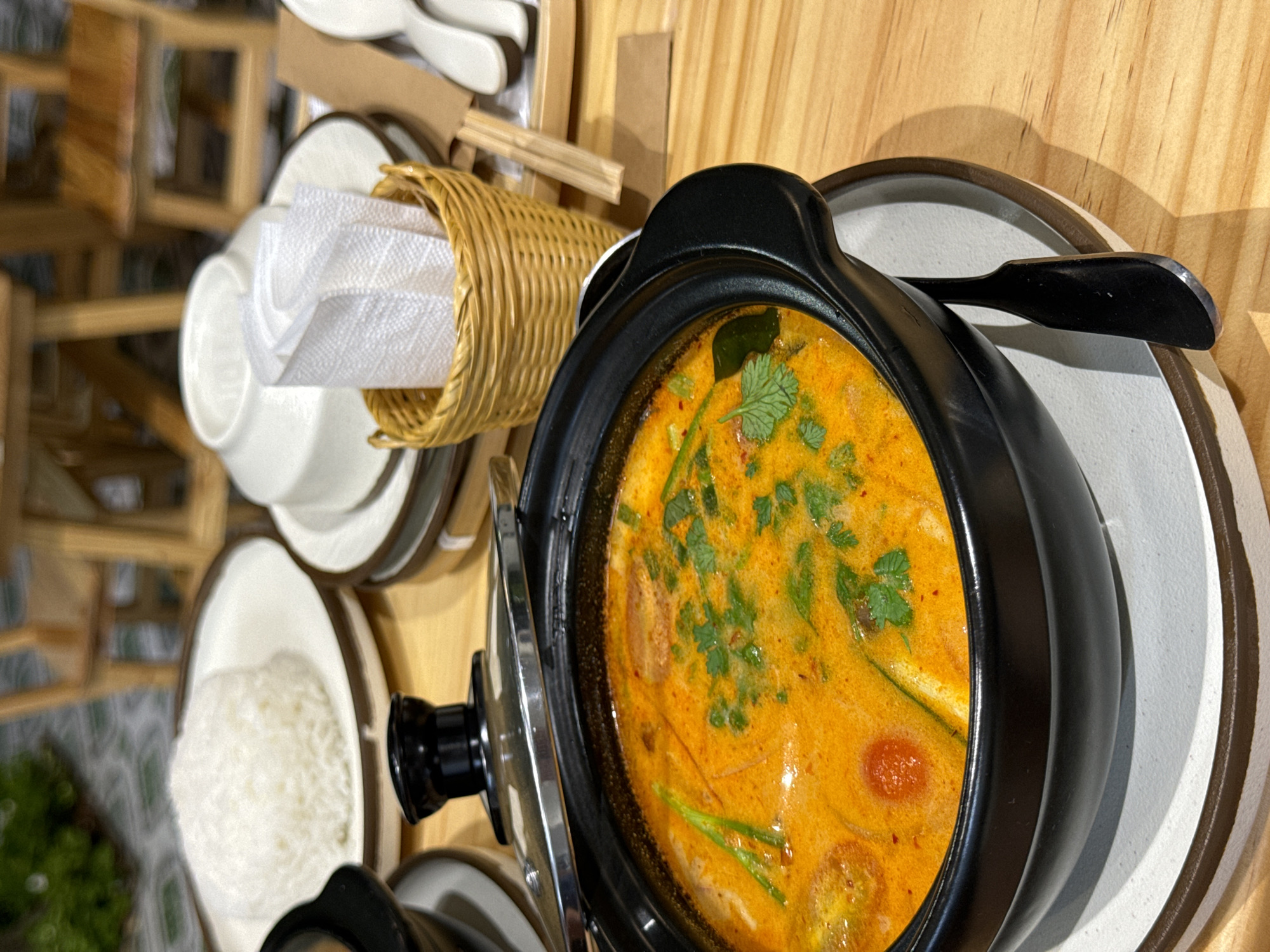 A close-up, high-angle shot shows a black pot filled with orange soup, garnished with green herbs. The pot rests on a plate. To the left, a small woven basket contains white napkins, next to stacked white bowls and plates. Further left, a bowl of rice is visible. The scene is set on a wooden table. The location is Phu Thuy Ward, Vietnam.