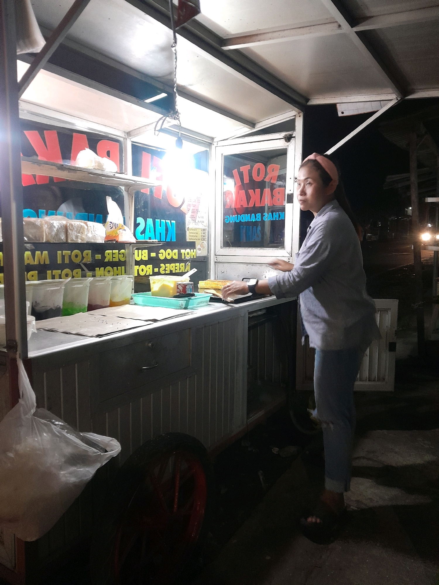 A woman stands behind a food cart at night in Tegalarum, Indonesia. The cart is illuminated by a bright light bulb hanging from the ceiling. The woman, wearing a light blue long-sleeved shirt and light blue pants, has a pink headband on. She is looking to her left, with her hands near the counter. The cart has a display of bread and various containers filled with colorful liquids, possibly ingredients for food. Signs on the cart are in Indonesian, advertising "ROTI BAKAR" (toast) and "KHAS BANDUNG" (specialty of Bandung). In the background, there are glimpses of other carts and dimly lit buildings, suggesting a street food scene. A large plastic bag hangs from the left side of the cart, and a red-rimmed wheel is visible on the lower left. The ground is paved with concrete.