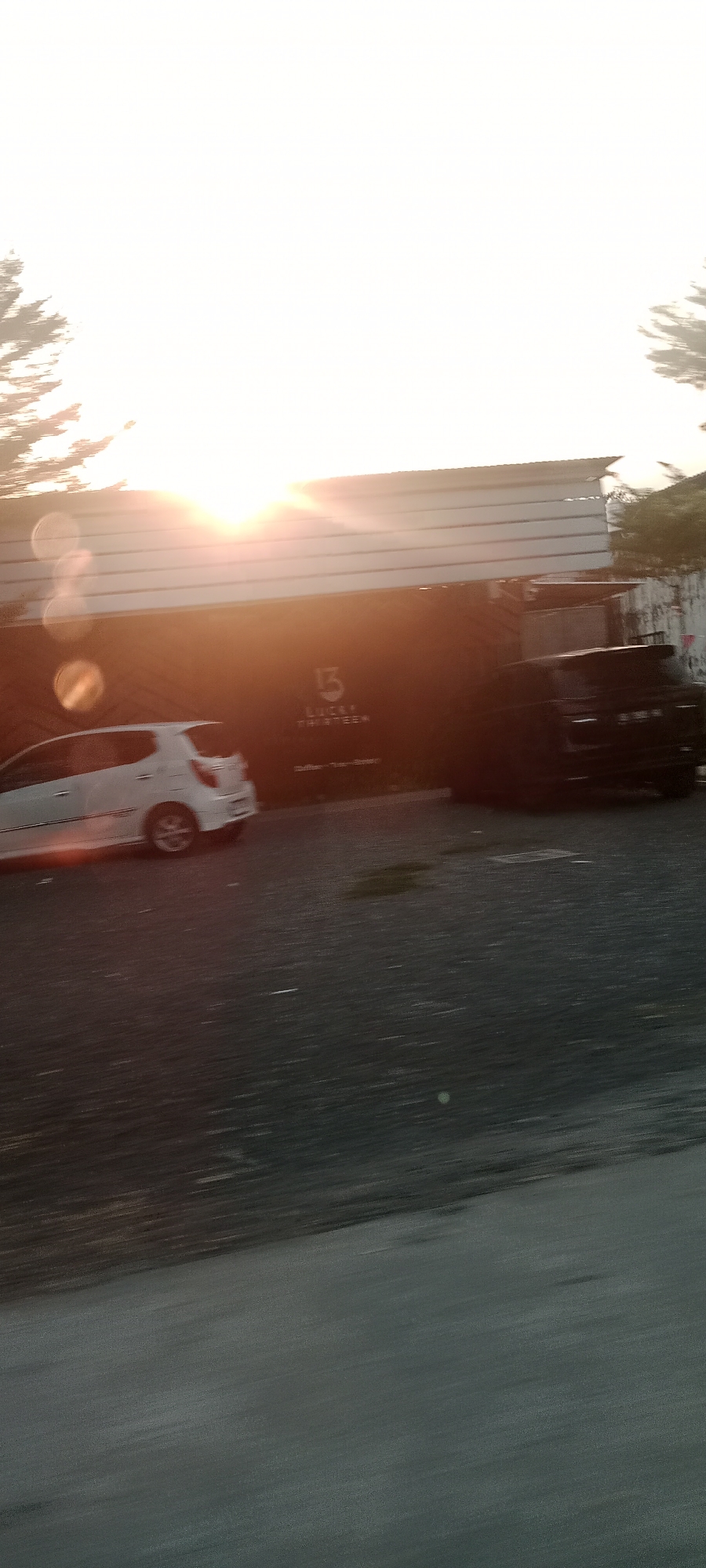 In Banyuraden, Indonesia, a white car is parked in front of a building with a dark facade. The building displays the "15 LUCKY THIRTEEN" logo in white lettering. To the right of the white car, a darker vehicle is partially visible. The sun is setting, casting a warm glow and lens flares across the scene. The ground in front of the building is paved.