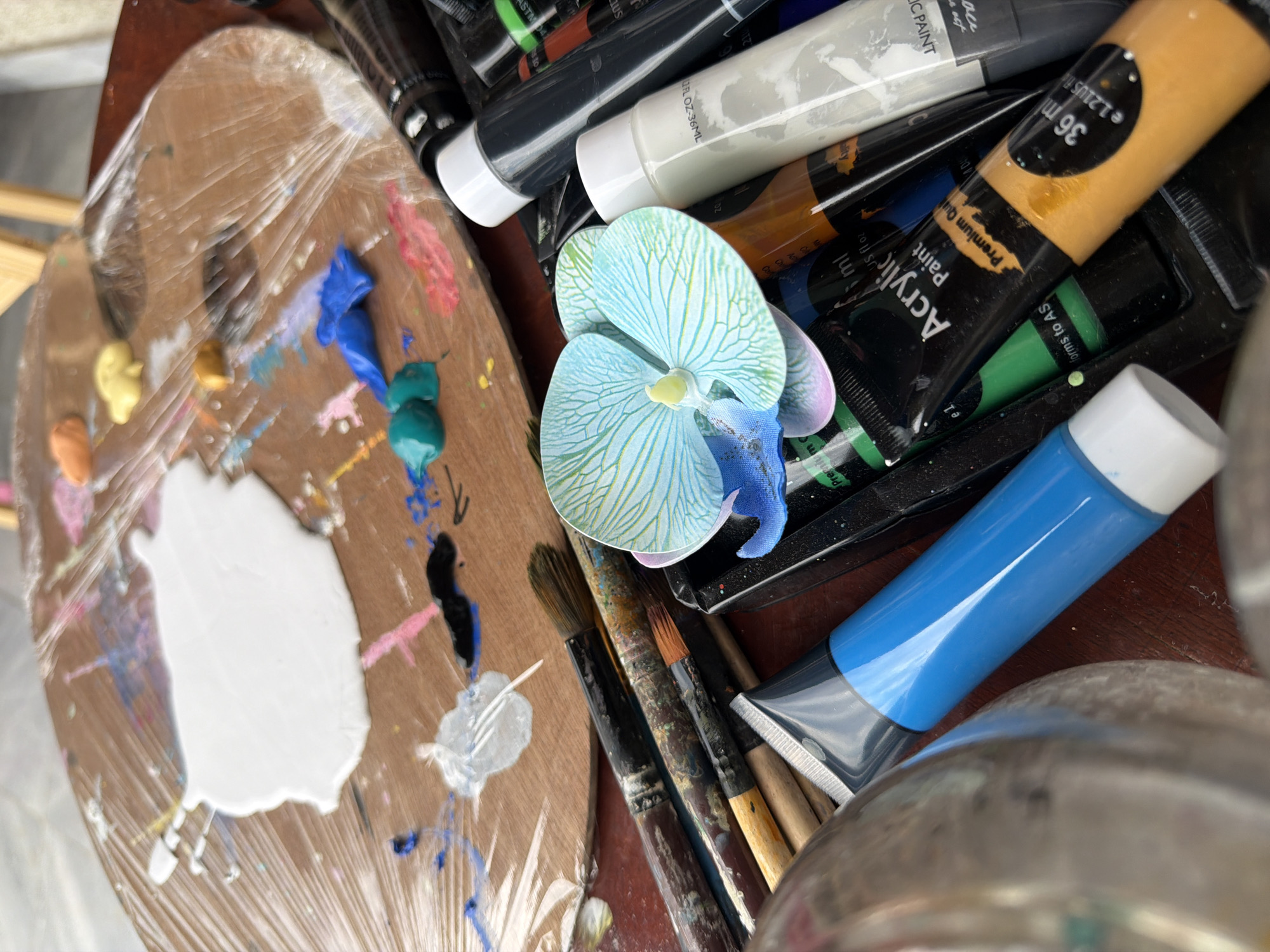 The image depicts an artist's workspace. A wooden palette, covered in plastic wrap, is adorned with various dollops of paint, including white, orange, yellow, teal, blue, and pink splatters. Several paint brushes, some appearing well-used and stained with paint, are arranged diagonally across the foreground. To the right of the palette, a collection of paint tubes is visible, including tubes labeled "Acrylic Paint". A light blue artificial orchid lies amongst the paint tubes. A metallic container is partially visible on the left side of the frame. The scene is set on a dark wooden surface. There is no indication of the city of Svilengrad, Bulgaria, or any people in the image.