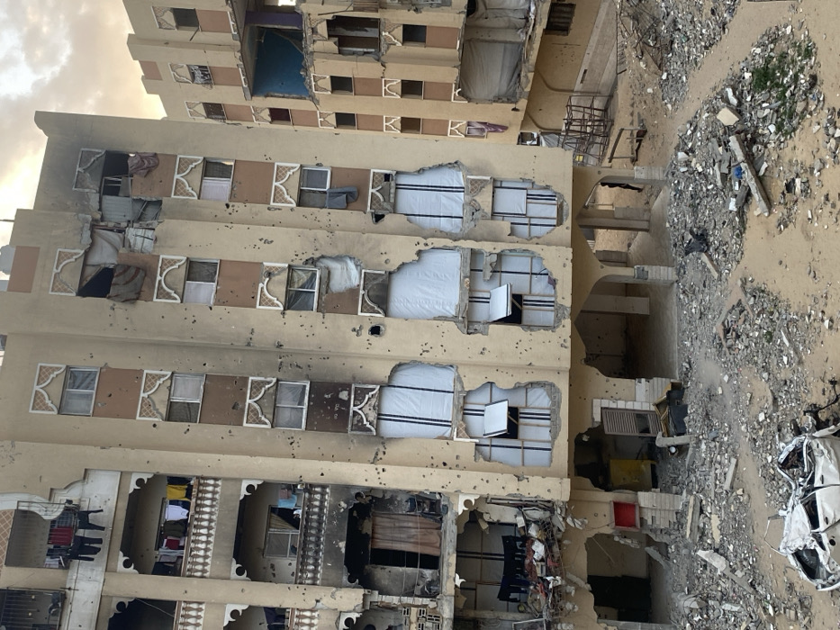 A multi-story building shows extensive damage, with gaping holes in its walls and missing windows. Bullet holes pepper the facade. Debris litters the ground surrounding the building, including a damaged car. Laundry hangs from some of the remaining balconies, suggesting recent habitation despite the destruction. The sky is overcast, lending a somber tone to the scene.