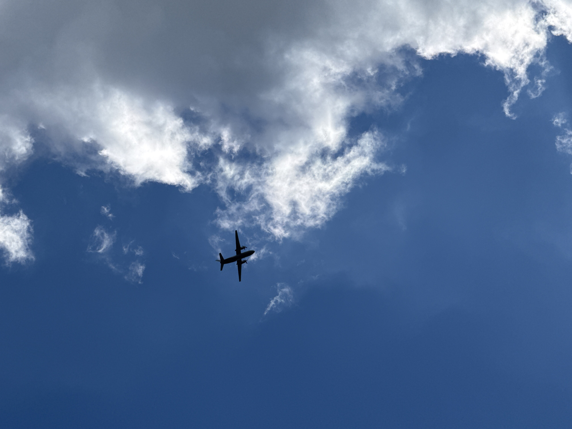 The image shows an aircraft silhouetted against a bright blue sky with some white clouds. The location is indicated as Rostov-on-Don, Russia. The aircraft is positioned in the lower center of the frame, its shape clearly defined against the brighter background. The sky occupies most of the frame, ranging from a deep blue to lighter shades where the clouds are. The clouds are mainly situated in the upper left portion of the image. There are no other discernible objects or people visible in the scene.