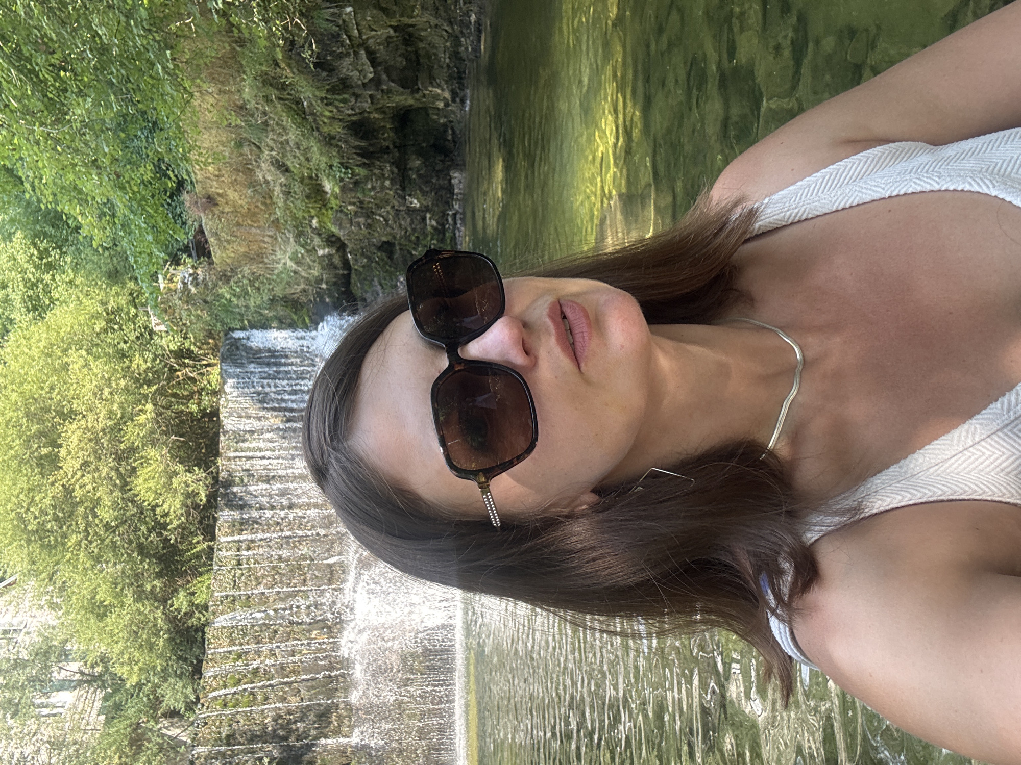 The media file is a selfie taken outdoors. The subject is a person with brown hair, wearing sunglasses and a white top with a textured pattern. They are in a natural setting with lush green trees and a waterfall or tiered stream in the background. The water is clear and reflects the sunlight. The scene appears to be tranquil and idyllic, possibly suggesting a warm and sunny day. The subject's face is partially visible, and their lips are parted as if speaking or about to speak. The overall mood of the image is relaxed and summery. The location is identified as Saint-Claude, France. There is no visible text in the image.