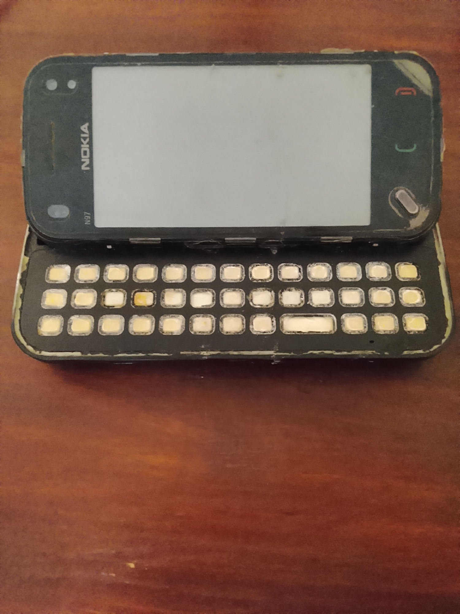 A Nokia N97 mobile phone is lying on a wooden surface. The phone is black and has a full QWERTY keyboard that slides out from underneath the screen. The screen is large and rectangular, and the Nokia logo is visible above it. The phone appears to be old and worn, with some scratches and scuffs on the casing. The wooden surface is a dark brown color and has a visible grain. The image is taken from a top-down perspective, and the lighting is somewhat dim. The location in Karangklesem, Indonesia, is not discernible from the image itself.