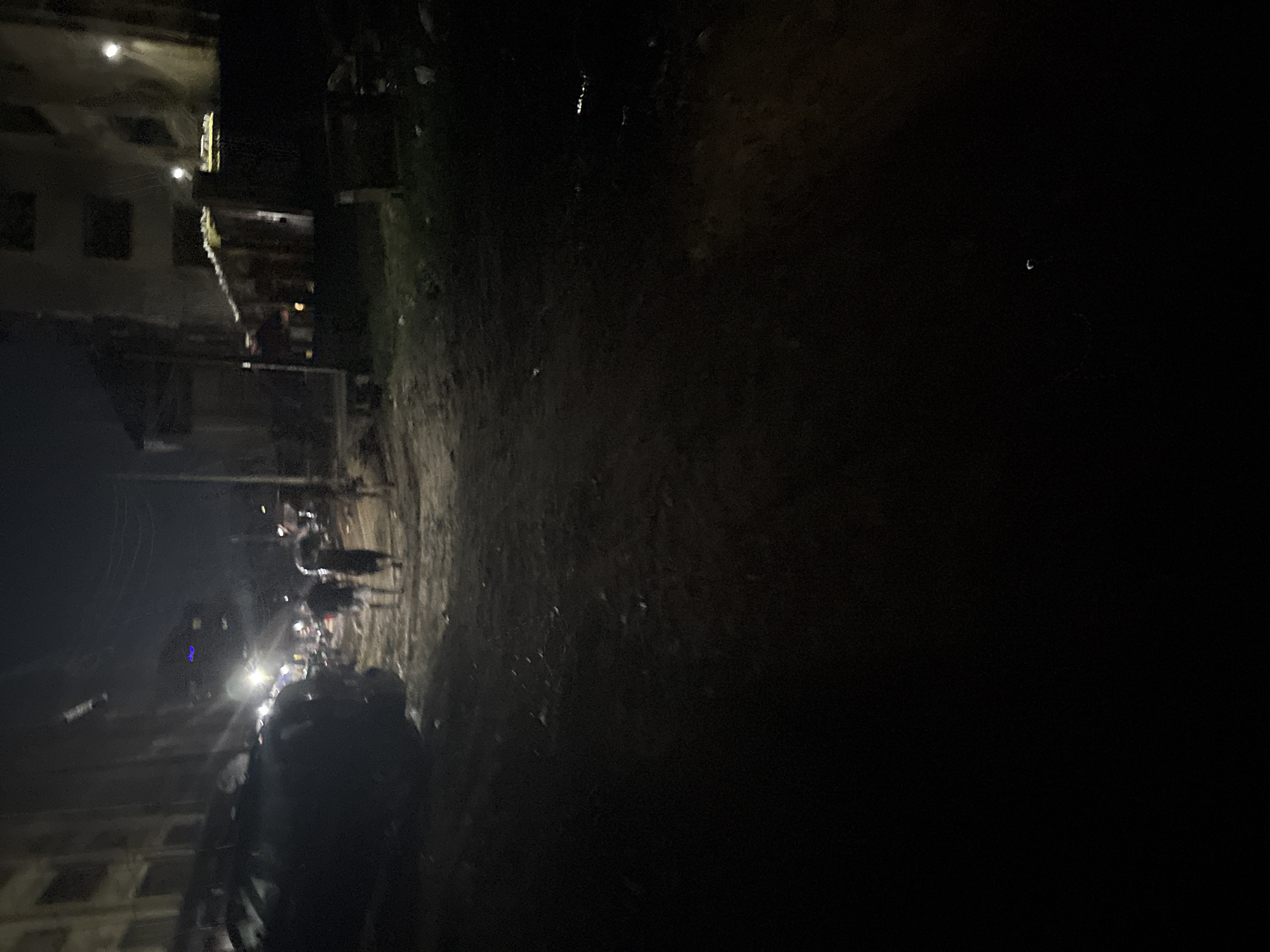 The image depicts a dimly lit street scene at night in Onitsha, Nigeria. A dirt road, littered with debris, occupies the foreground and middle ground. To the left, a dark-colored vehicle is partially visible, parked alongside buildings. In the middle distance, three individuals are walking away from the camera, their forms indistinct in the low light. They appear to be casually strolling down the street. Further back, buildings line both sides of the road, with some windows illuminated, suggesting interior lighting. A small, makeshift structure with a corrugated roof is also present. The overall atmosphere is one of a typical, somewhat unlit, urban environment at night in Nigeria.