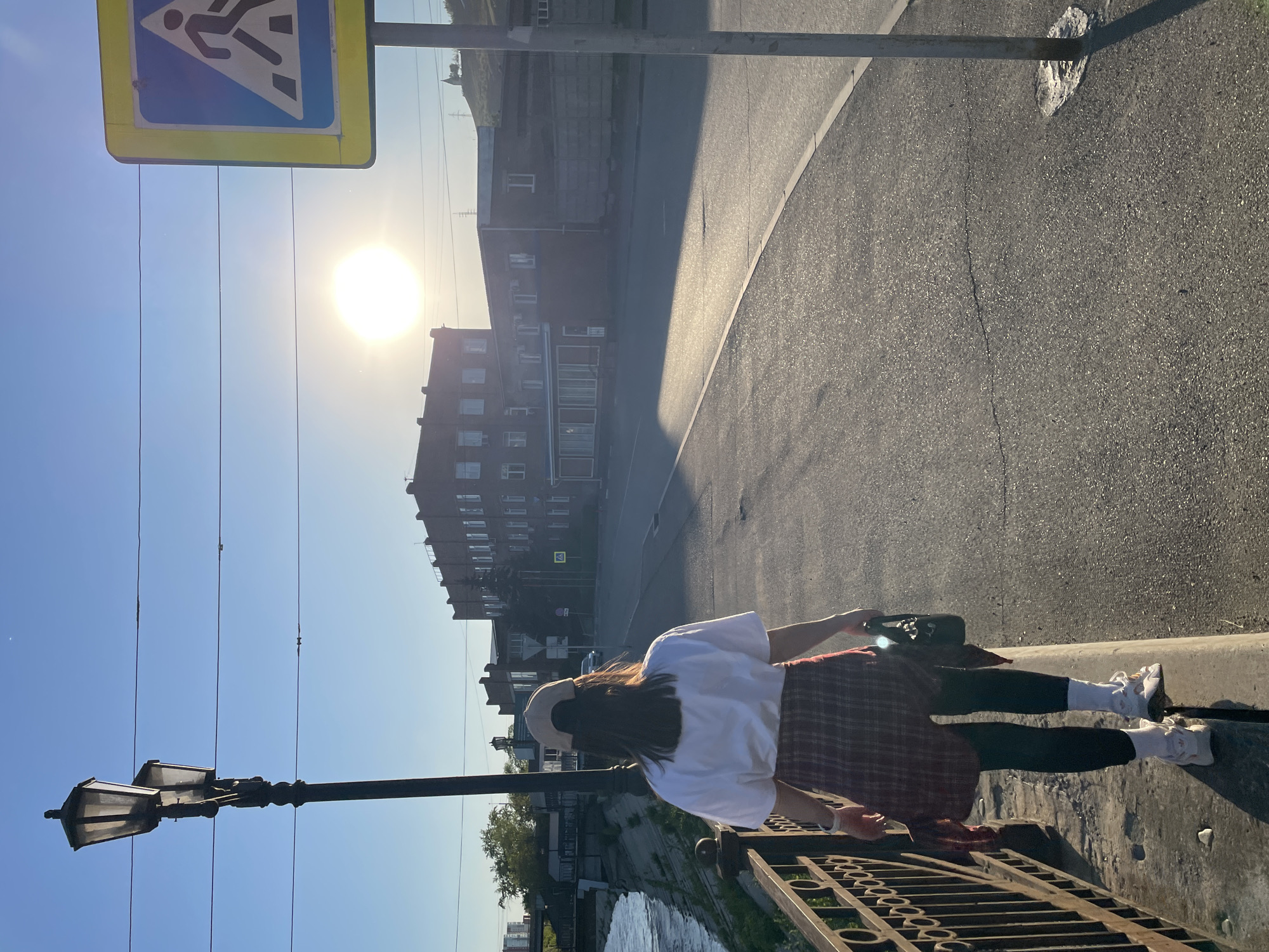 A medium shot shows a woman standing on a bridge in Krasnoyarsk, Russia, facing a street with buildings in the background on a sunny day. The woman has long dark hair, wears a white shirt, a plaid skirt, black leggings, white sneakers, and a light-colored hat. She holds a black bag in her left hand and rests her hands on a dark metal railing. Behind the railing is a river with green foliage on the riverbank. To the left, a lamp post and overhead wires are visible against a blue sky. To the right is an asphalt street that curves slightly, with a building on the other side and a yellow pedestrian crossing sign is visible above the lamp post. The sun is high in the sky, creating a strong glare.