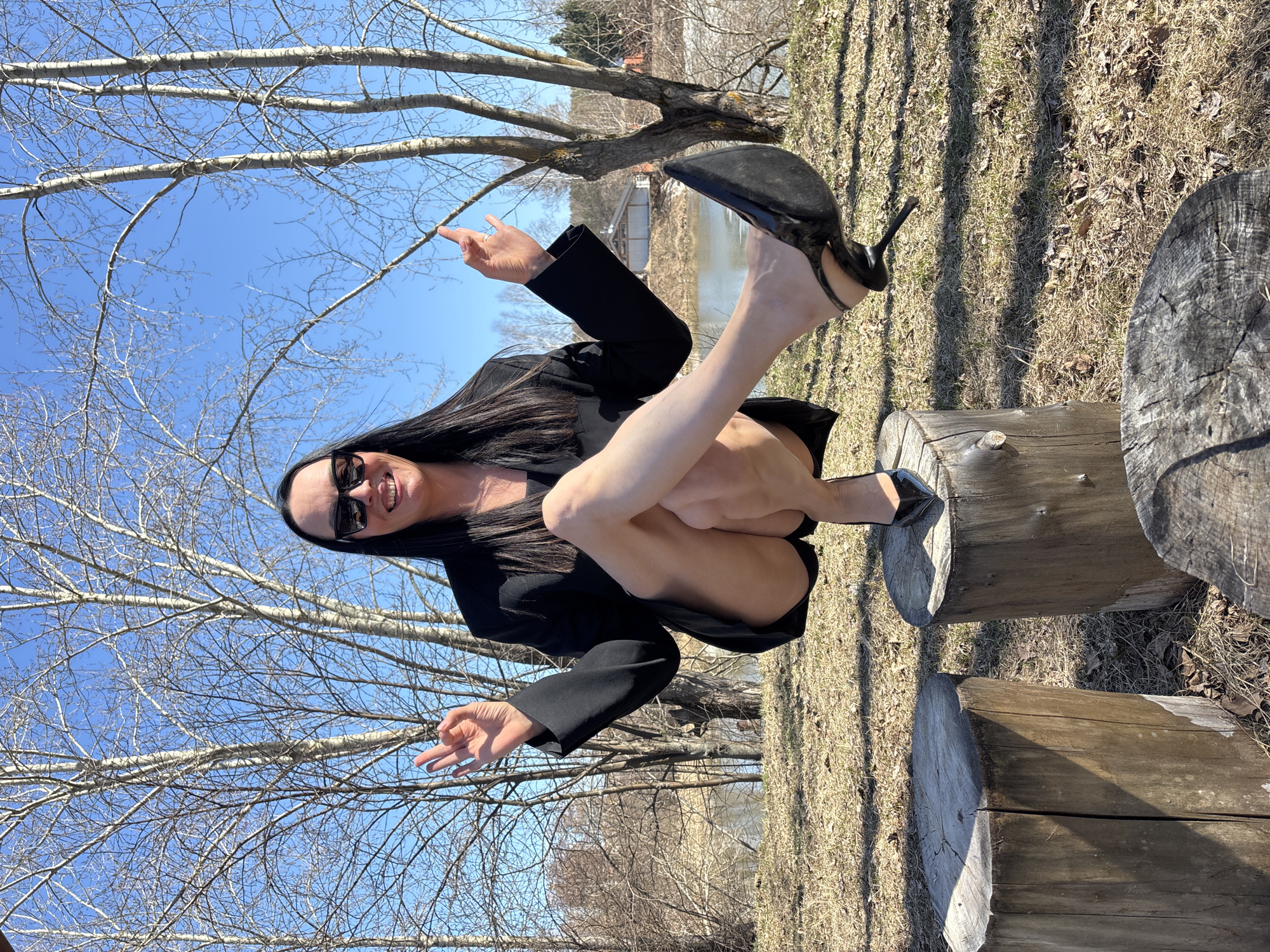 A woman sits on a cut log in a park in Chaikovsky, Russia, wearing a black blazer, dark sunglasses, and high heels. She points upward with her right hand and makes a circle with her thumb and index finger on her left hand. Bare trees stand in the background under a clear blue sky. Dry grass covers the ground. A body of water is visible through the trees.