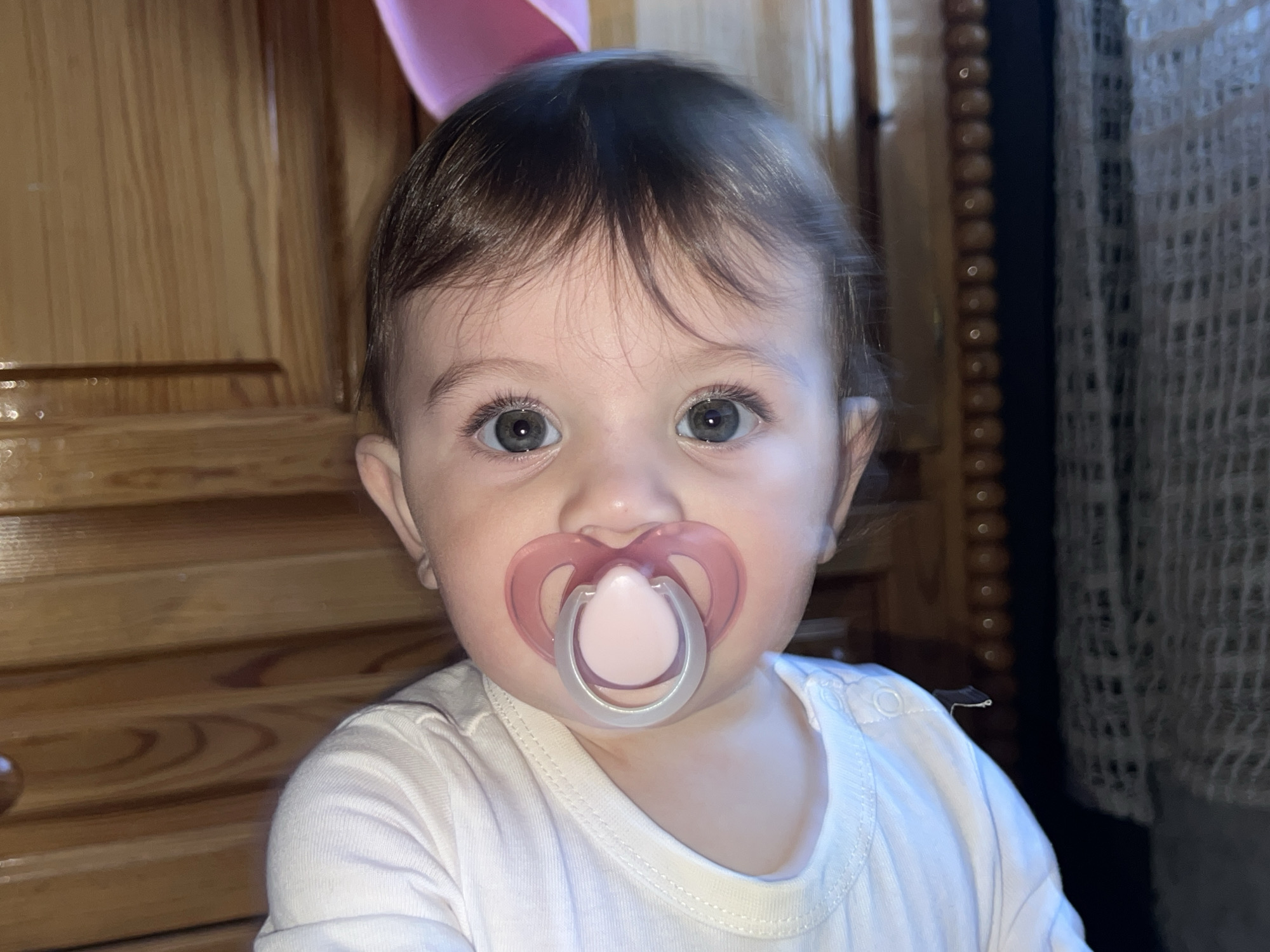 An infant is depicted in a close-up, frontal portrait, indoors in Elena, Bulgaria. The subject is a young child with fair skin, dark brown hair, and blue-grey eyes, looking directly at the camera. A pink and white pacifier is positioned in the infant's mouth. The infant is wearing a plain white, long-sleeved shirt with visible texture on the right shoulder. The background to the left features a vertical wooden surface, possibly a door or wall paneling, with horizontal grain. A partial view of a pink balloon is visible in the upper left. To the right, a darker vertical element with a decorative beaded trim separates the wooden surface from a light-colored, loosely woven or crocheted curtain. The lighting suggests an indoor, possibly low-light environment.