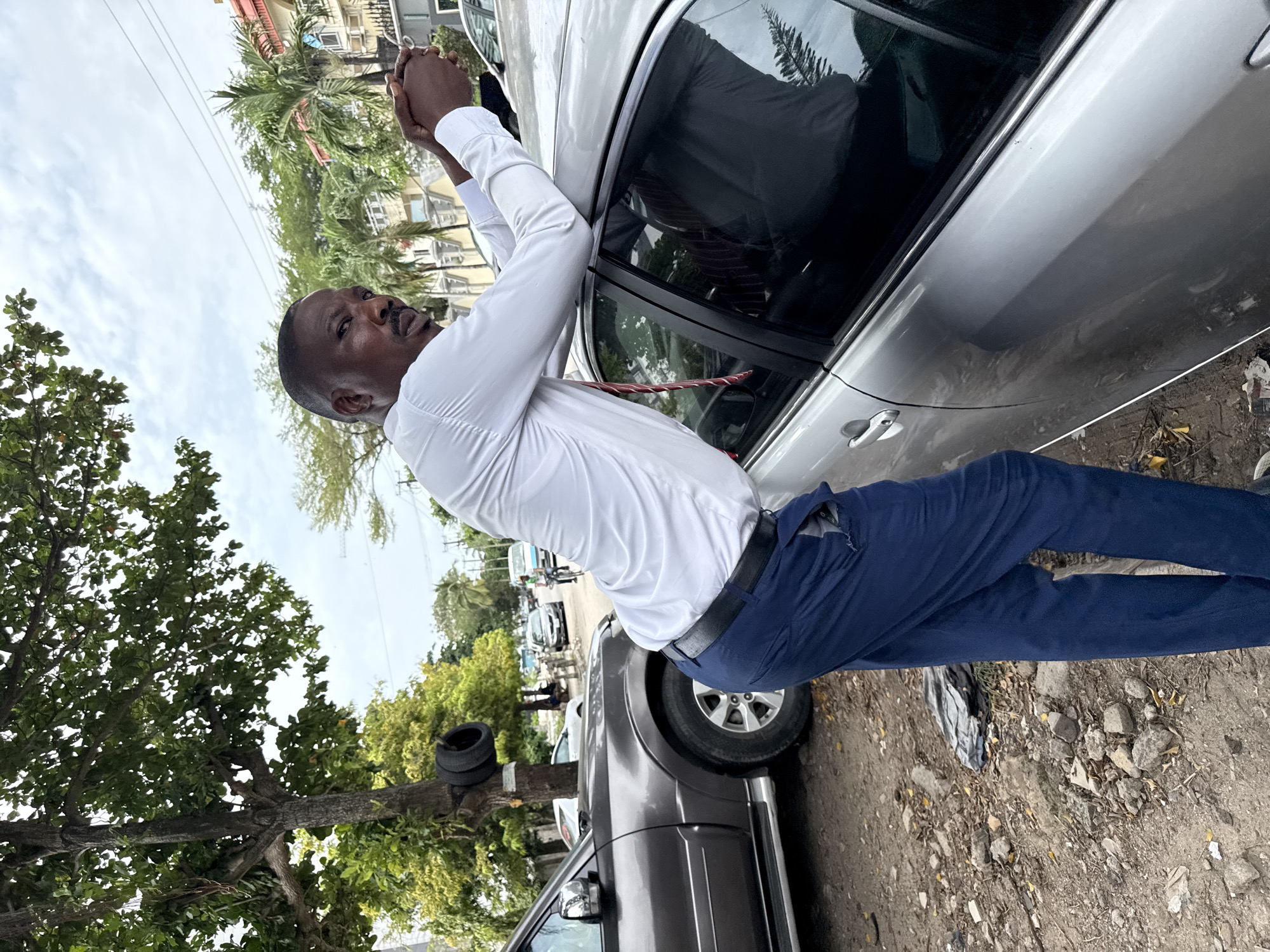 A man in a white shirt and blue pants stands next to a silver sedan in Itirin, Nigeria. He is holding the top of the car door frame with both hands. A red and white striped object is hanging from his neck. The ground is dirt and gravel, with trash and debris. In the background are other cars, buildings, and trees, including one tree with two tires attached to it. Power lines are visible overhead.
