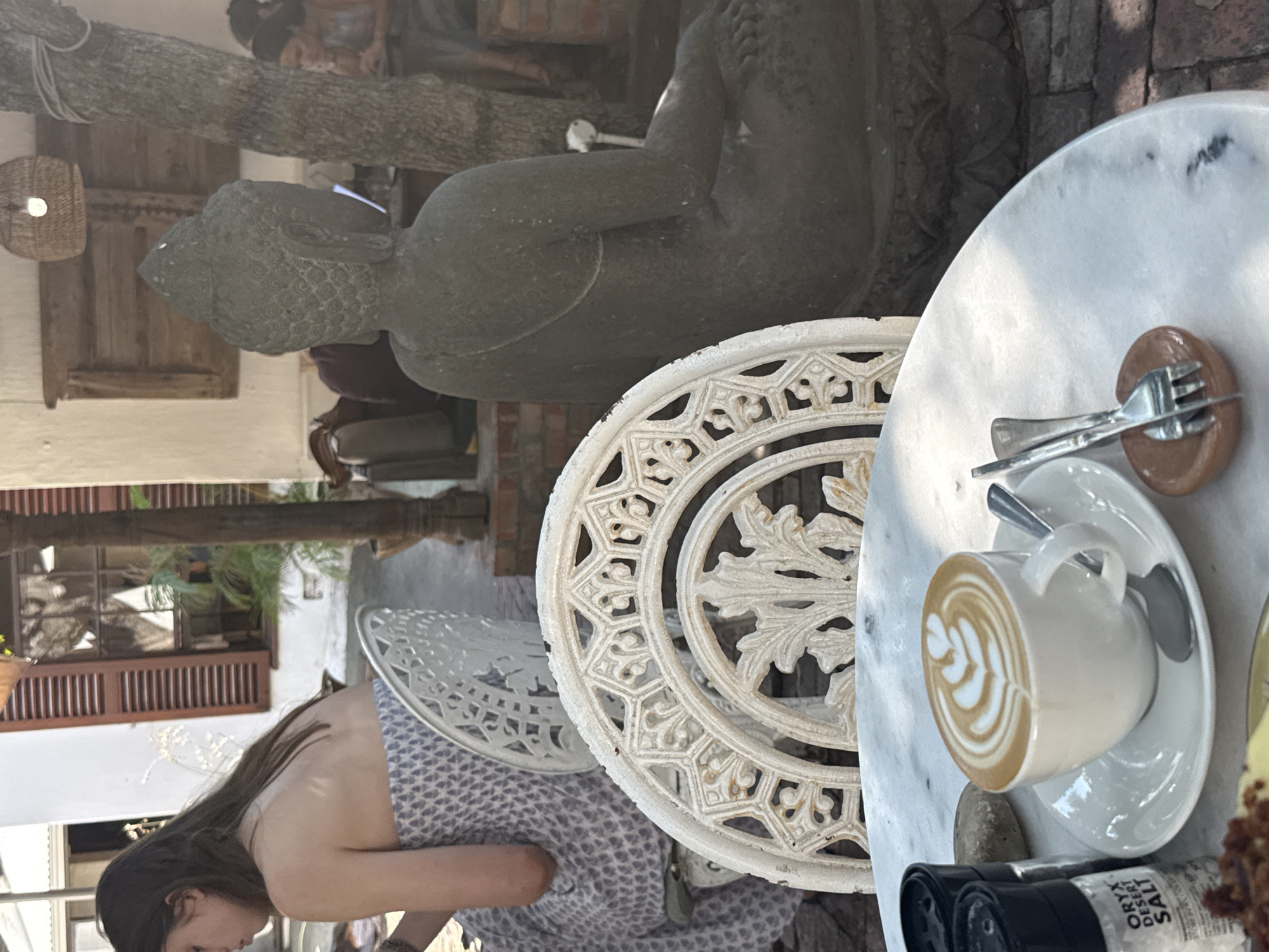 The image captures a cafe scene in Cape Town, South Africa. A woman with long brown hair sits at a white, ornately designed metal chair and table. On the marble-topped table, there's a white cup of coffee with latte art on a saucer, alongside silverware on a small plate and condiments. Behind the chair is a stone Buddha statue, positioned near a tree. In the background, other patrons are visible, adding to the cafe ambiance. The architecture includes wooden doors and windows, suggesting a historic building. Overall, the scene reflects a relaxed outdoor cafe setting.