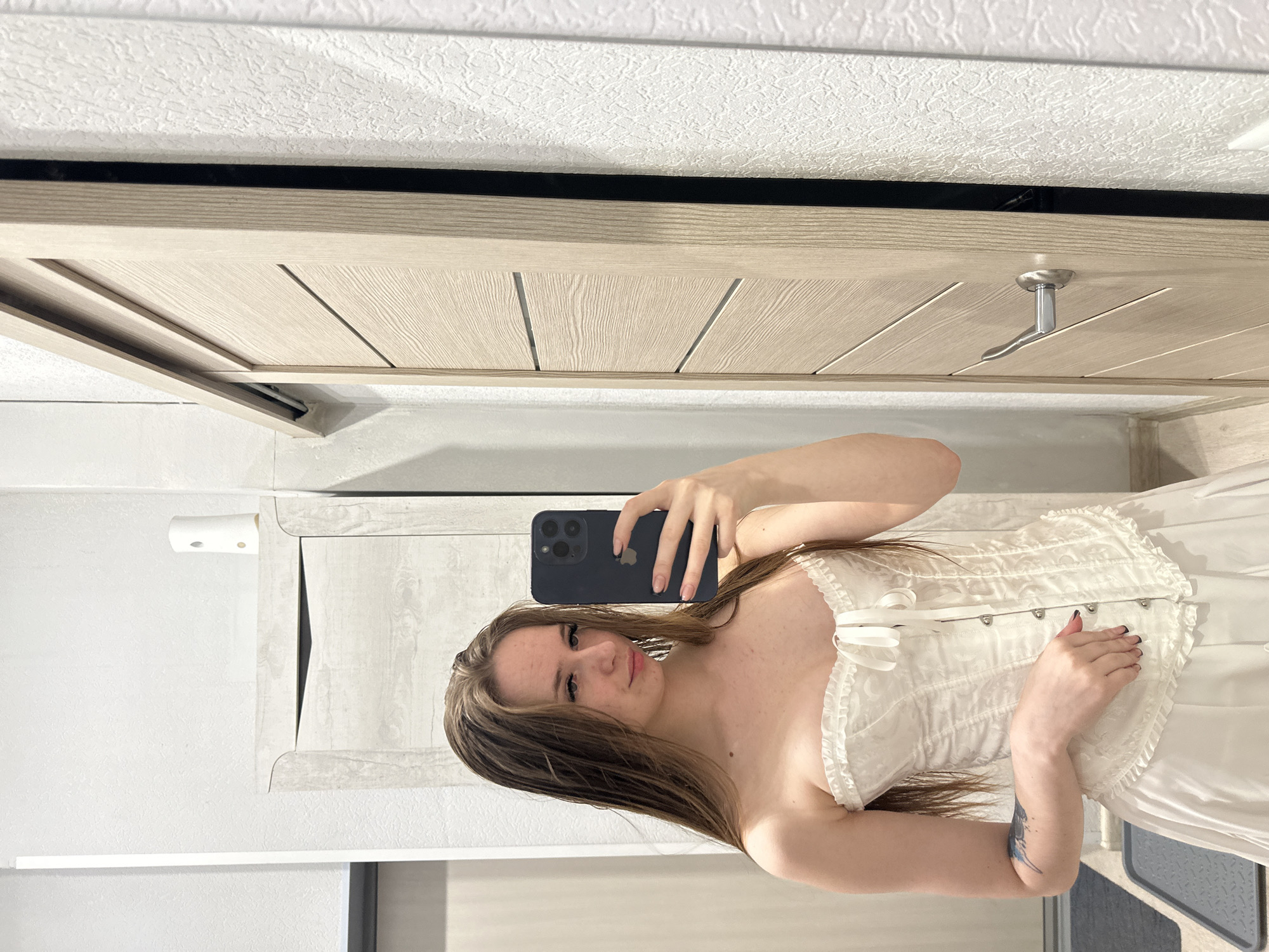 A young woman is taking a selfie in what appears to be a bathroom or bedroom setting. She is wearing a white corset and a white skirt or dress. Her hair is long and light brown. She is holding a dark-colored smartphone, obscuring part of her face. The background shows a door with a modern design, possibly made of light-colored wood, and a wall painted in a pale color. A white object, possibly a dispenser, is mounted on the wall. The overall lighting is bright and artificial. The setting appears to be in a private residence, but the location is Unknown, Unknown.