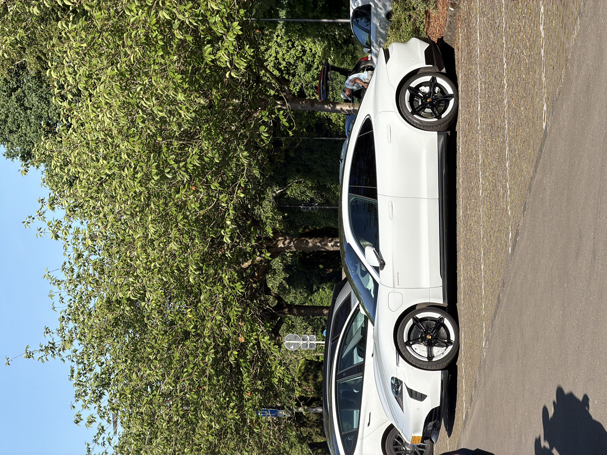 The image shows a parking area in Fentange, Luxembourg, with two white Porsche Taycan electric vehicles parked adjacent to each other. A large green tree is present on the left side of the frame, partially obscuring the sky. In the background, there are more parked cars and a person is visible near a car. The parking area is paved, and marked parking lines are present.