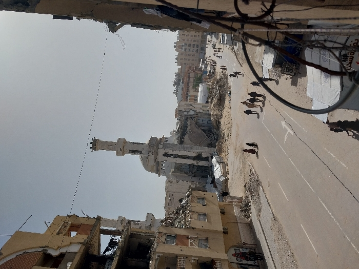 An aerial view from an elevated structure reveals a heavily damaged urban street scene in Khan Yunis, Palestinian Territory, under a clear, bright sky.
In the mid-ground, a large mosque, identifiable by its tall, cylindrical minaret with a crenellated top and a prominent central dome, stands significantly damaged. Extensive sections of the mosque's structure appear collapsed or missing.
To the left of the street, a multi-story building is severely compromised, with exposed interiors, shattered walls, and visible structural damage. Piles of rubble and debris surround its base and extend into the foreground. On the immediate right, the structure from which the image is taken also shows signs of damage, with exposed wiring and architectural elements in the foreground.
The central paved road features faded white lane markings and a distinct white turn arrow. Numerous individuals are seen walking along the street and its shoulders. In the left-midground, a group of approximately four people, including one pushing a stroller or small cart, moves along the road. Another cluster of about six people walks further down the street on the right. Scattered individuals are visible moving in various directions towards the background.
Extensive debris fields and remnants of destroyed buildings flank both sides of the road beyond the immediate structures. In the distant background, additional multi-story buildings are visible, some appearing less damaged. Patches of white tarpaulin or makeshift shelters are visible near the right foreground, against the damaged buildings.