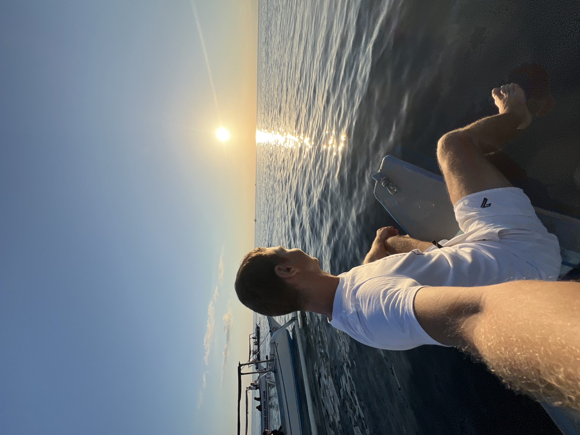 A man with short brown hair, wearing a white t-shirt and white shorts, is sitting on a boat and looking out at the ocean. The sun is setting on the horizon, casting a reflection on the water. There are other boats in the distance. The man's arm is in the foreground, and his leg is visible, with his foot resting on the boat. The location is Unknown, Indonesia.