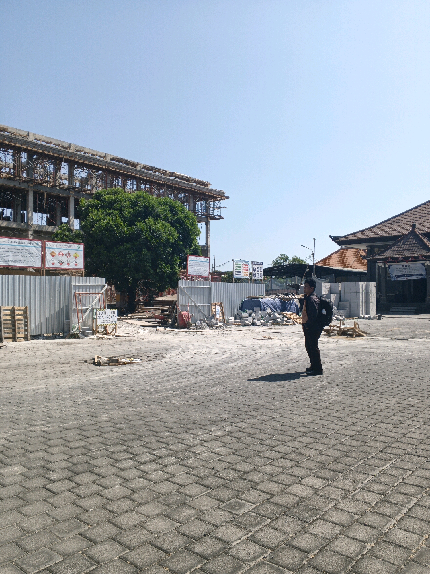 An outdoor, low-angle, full-shot image shows a man with a backpack standing in a paved area in front of a construction site and a building with a traditional roof. The man is facing the right of the frame, with his left shoulder slightly forward. He is wearing a dark jacket over a light-colored shirt and dark pants. His backpack is black and appears to be of moderate size. The paved area is made of interlocking rectangular concrete blocks, and the man's shadow is cast to his left.
In the background, a multi-story building is under construction, with visible scaffolding and concrete structures. A large, lush green tree stands near the construction site. To the right of the construction, a building with an orange-tiled, pitched roof and dark wooden eaves is visible. This building has an entrance with steps. Various construction materials are scattered around the site, including wooden pallets, piles of concrete blocks, and tarpaulin-covered objects. Several signs are posted on the construction fence. The sky is clear and blue. The scene suggests an active construction project in an urban environment. Given the traditional roof style of the building and the presence of construction, this location could be an area undergoing development or renovation within Denpasar, Indonesia.