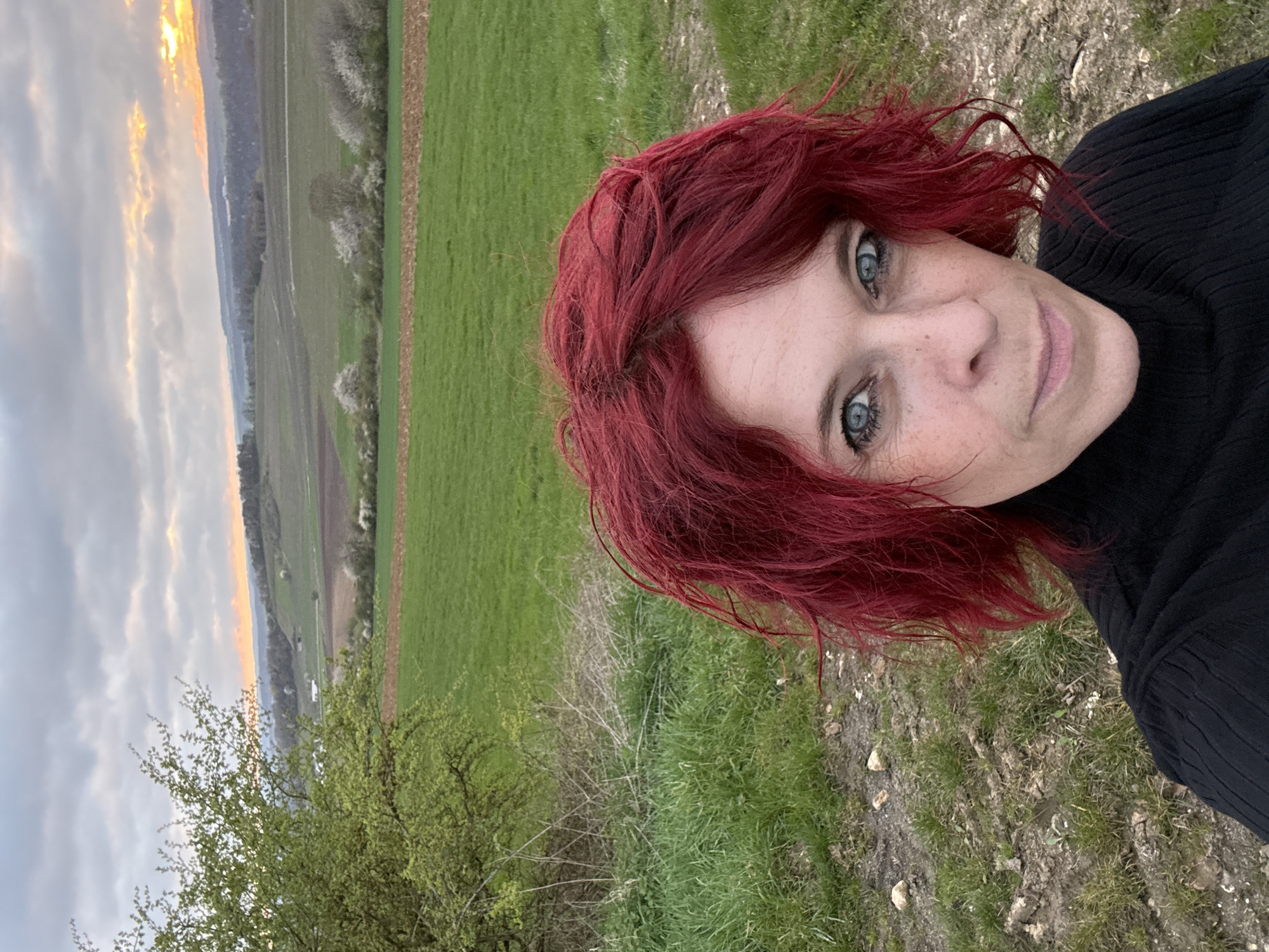 A medium shot captures a woman with shoulder-length, vibrant red curly hair against a landscape background. The woman has light skin, freckles, and striking blue eyes. She wears a black, textured top with a high neck. The background consists of a green field transitioning into a valley under an overcast sky with a touch of sunset glow on the horizon. A small tree with light green leaves is visible in the foreground. The ground around her appears to be grassy with patches of exposed soil. The location is not identifiable and is specified as Unknown, Unknown.