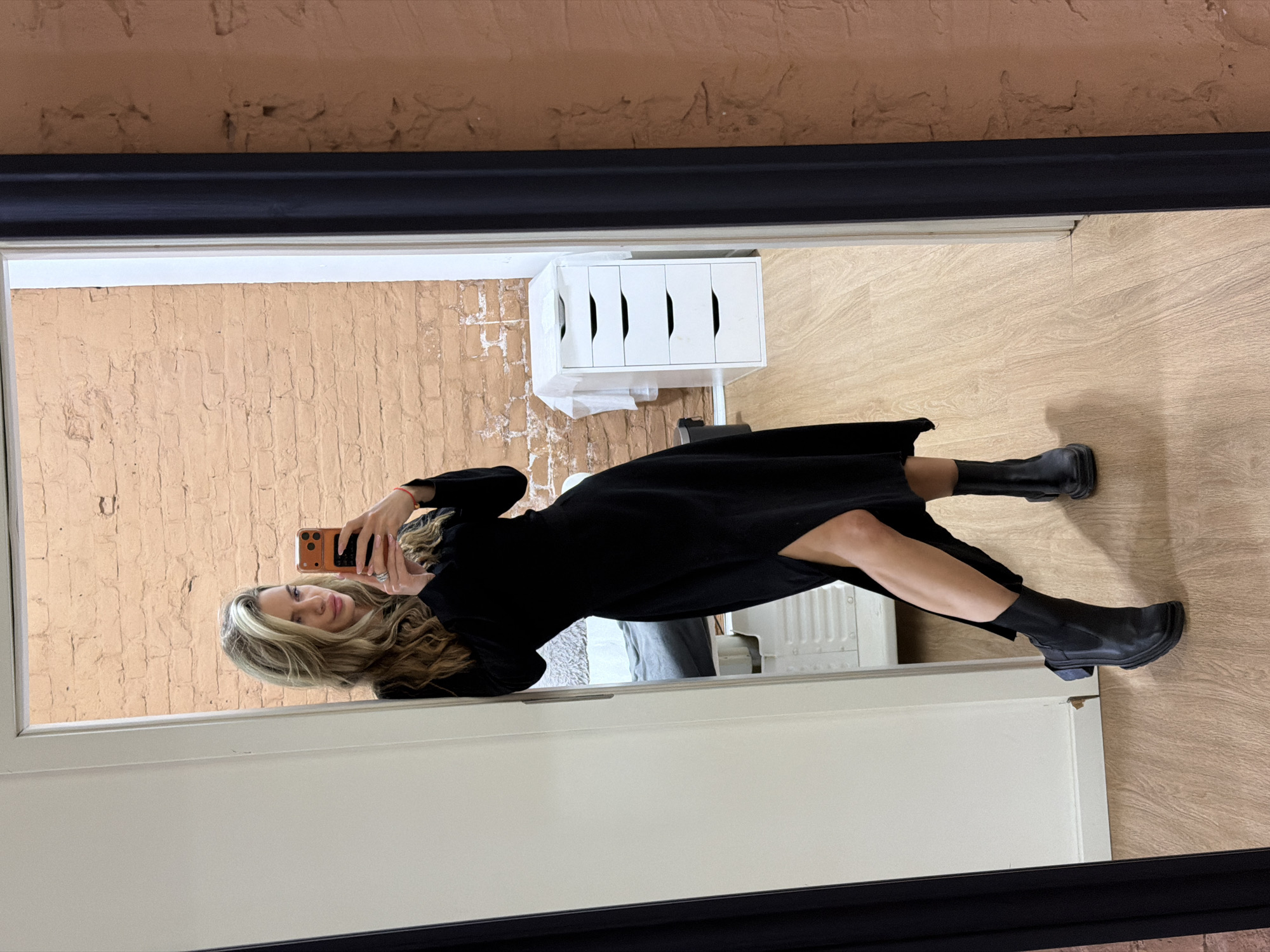 The image shows a woman reflected in a mirror. The woman has blonde hair and is wearing a black dress and black boots. She is holding a phone and taking a picture of herself in the mirror. The background of the image includes a brick wall, a white dresser, and a wooden floor. There is no information to indicate the location of the image is in Berlin, Germany.