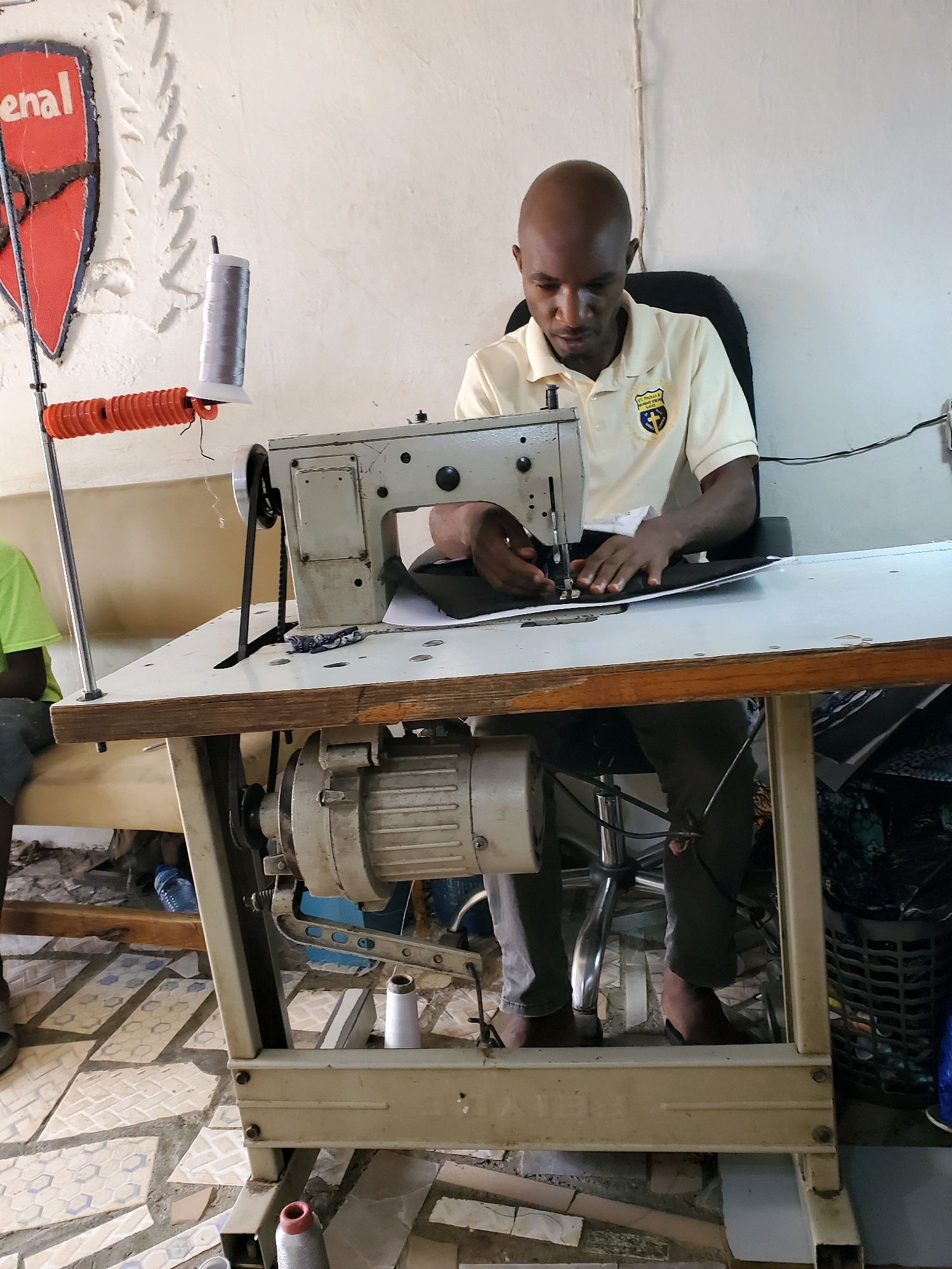 A bald Black man in a light yellow polo shirt is sitting at a sewing machine and sewing black fabric. The sewing machine is on a light-colored table with a wooden edge. The man is wearing grey pants. To the left of the man, a metal pole holds an orange object and a spool of grey thread. A person wearing a green shirt is visible in the background to the left. The floor is tiled with a patterned design. A large motor is attached to the sewing machine. There are spools of thread on the floor. A dark basket with a floral pattern is in the background to the right. A red and blue shield with the word "enal" is visible on the wall behind the man.