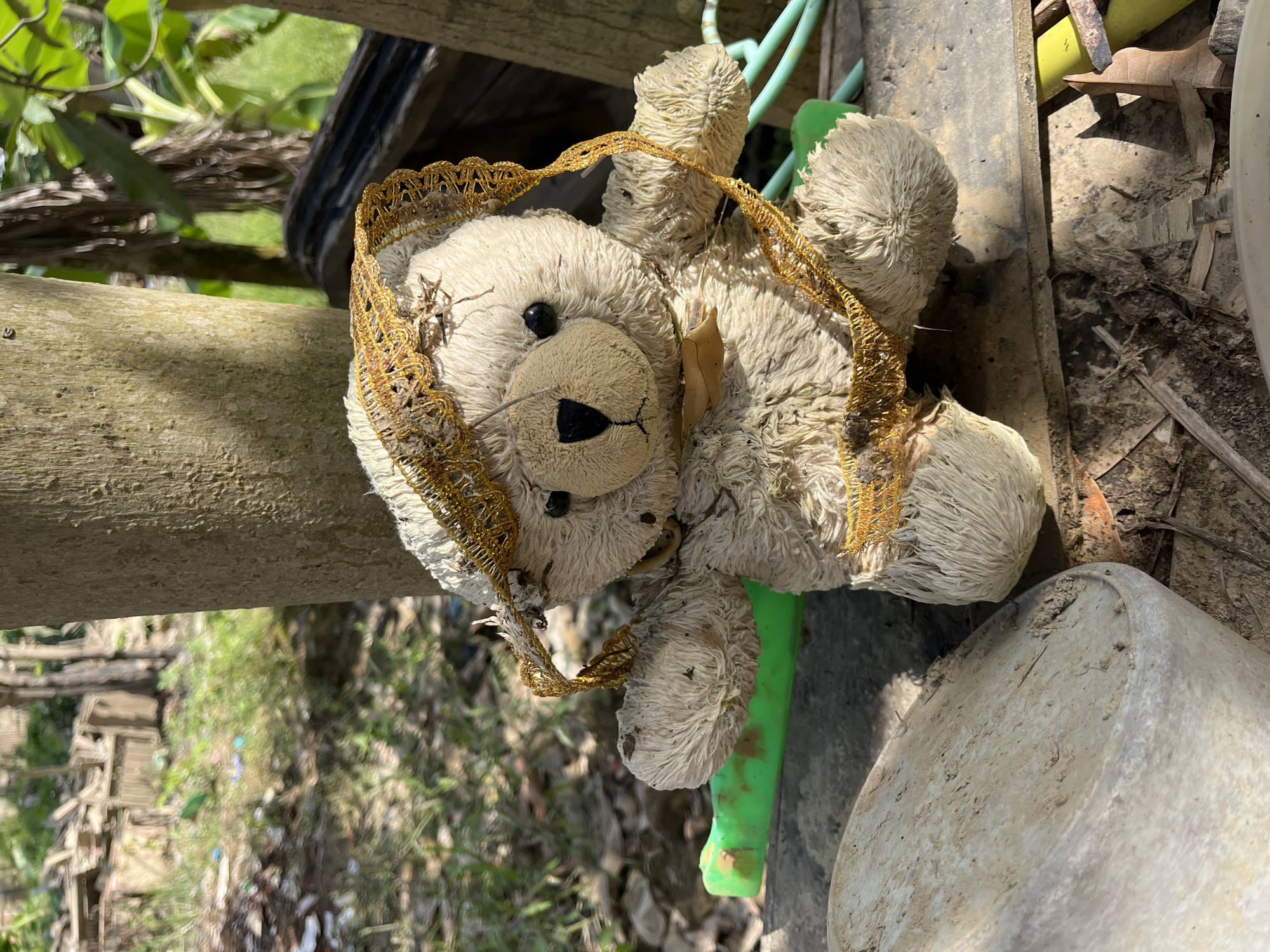 A solitary, weathered teddy bear is the focal point of this image, positioned outdoors amidst debris and vegetation. The bear, predominantly off-white and appearing aged and somewhat disheveled, wears a tattered gold ribbon or trim around its head and neck. Its fur is matted, and there are visible stains and dirt marks, suggesting exposure to the elements. The bear is seated on a rough, greyish surface, possibly wood or concrete, with a bright green object partially visible beneath its left arm.
The background suggests an unkempt outdoor environment. To the left, there's a blurry expanse of green grass interspersed with dried leaves, twigs, and what appear to be pieces of discarded material, including some colored plastic fragments. Further in the background, structures made of wood are faintly visible, along with trees and dense foliage, hinting at a somewhat overgrown or neglected area. To the right, parts of wooden structures and more greenery are present. Sunlight casts distinct shadows, indicating it is daytime. The scene evokes a sense of abandonment or disuse. The location is identified as Kota Kuala Simpang, Indonesia, a detail that places the scene within a specific geographical and cultural context, though the image itself does not contain explicit markers of this location beyond the provided information.
