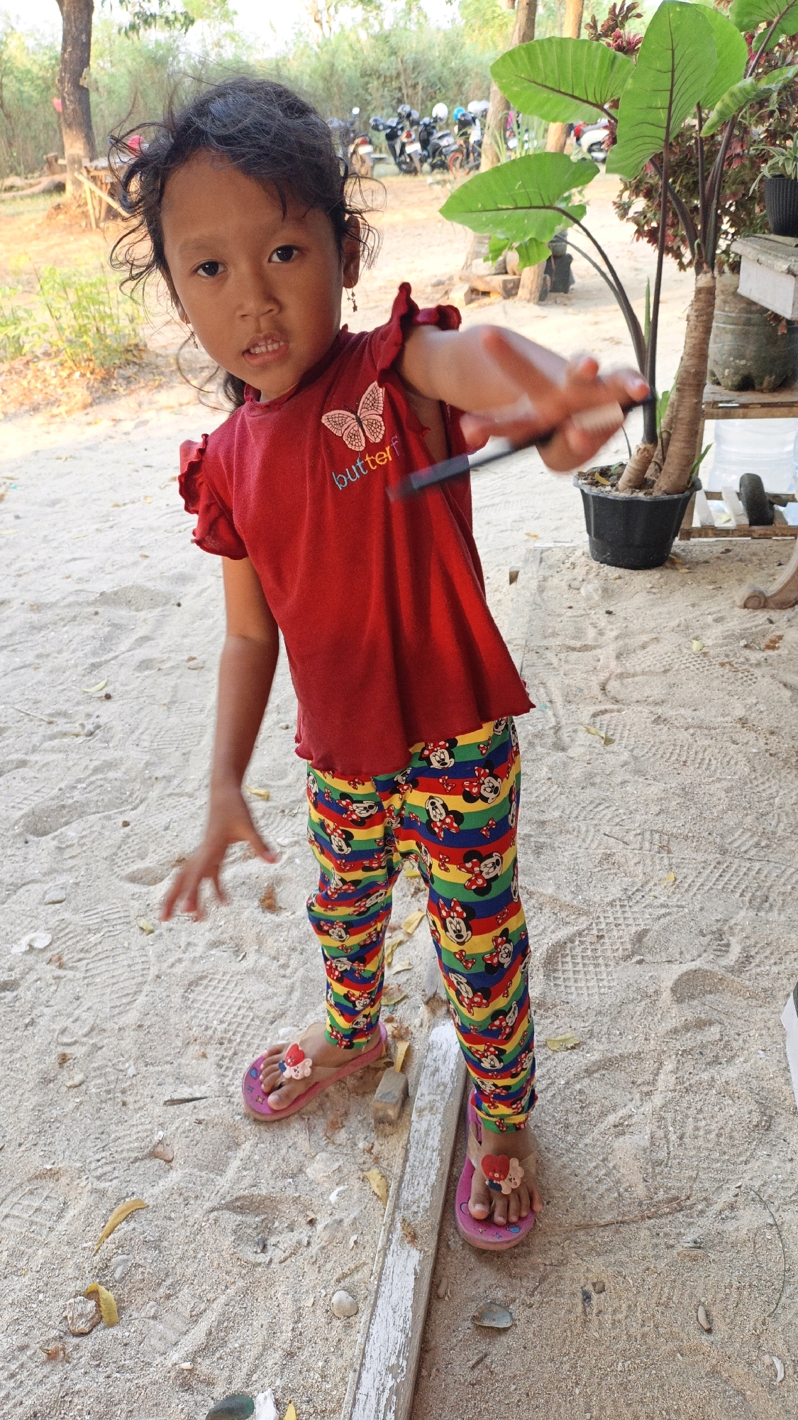 A young girl, appearing to be around 4-5 years old, stands on a sandy surface. She has dark, curly hair and is wearing a red t-shirt with a butterfly graphic and the word "butterf" printed on it. Her pants are colorful, striped with red, yellow, green, and blue, and feature a repeating pattern of Mickey Mouse characters. She is wearing pink flip-flops with floral embellishments. The girl is extending her right arm forward, holding a dark object that appears to be a pen or stylus. Her left arm is bent, with her hand open. In the background, there are trees, some motorcycles parked, and a large potted plant with broad leaves. The scene suggests an outdoor location, possibly a park or a beachside area in Cikal, Indonesia.