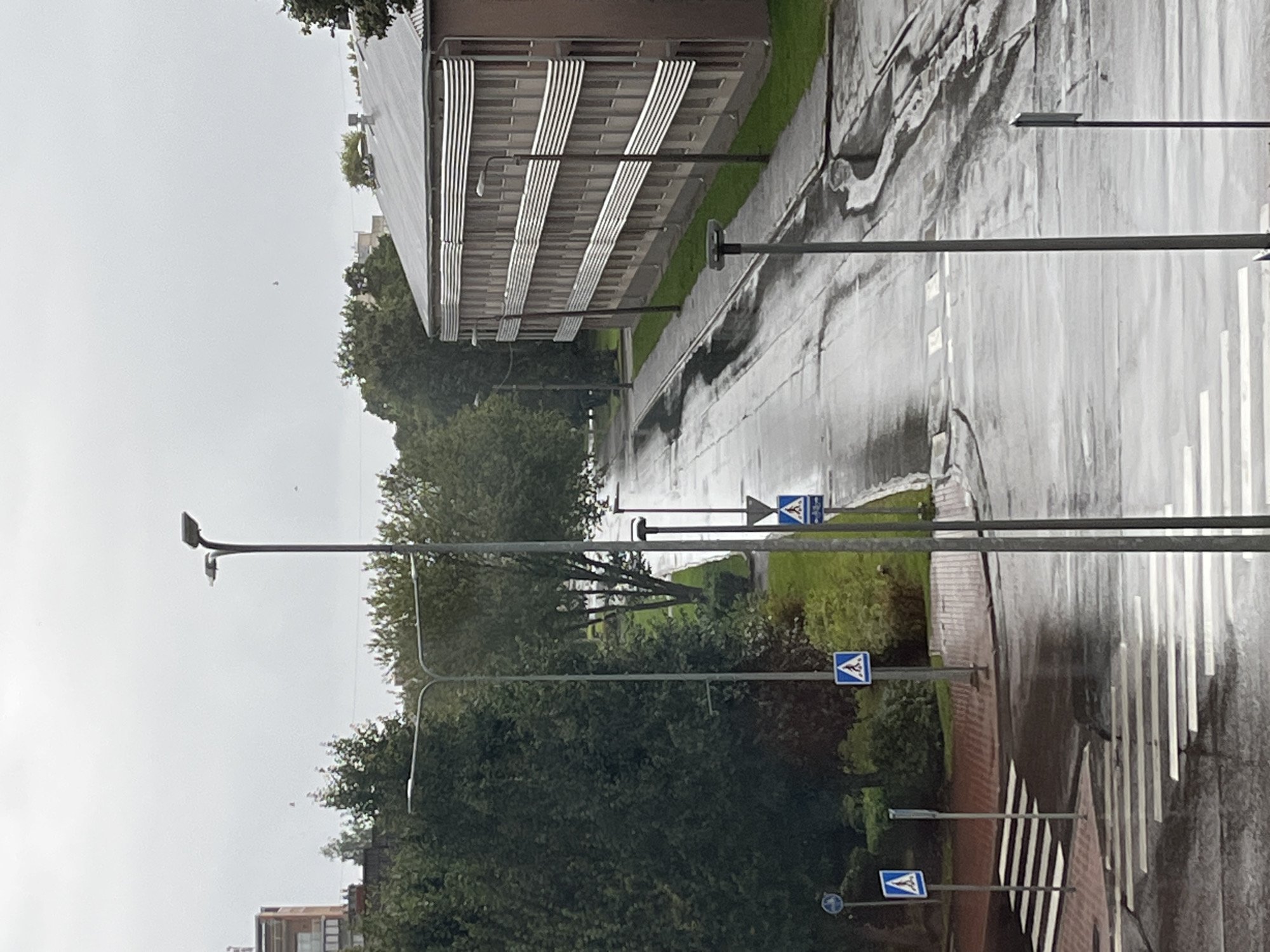 A wet street in Kohtla-Järve linn, Estonia, is captured on an overcast day. The road, reflecting the muted light, shows signs of recent rain with puddles and streaks across its surface. Multiple pedestrian crossings are visible, painted in bold white stripes, indicating a need for caution. Several streetlights stand along the pavement, their designs modern and functional. To the right, a multi-story building with a distinct facade, featuring horizontal metallic elements, dominates the scene. Trees and green grass line the sides of the road and the building, providing a natural contrast to the urban architecture. Traffic signs, including pedestrian crossing symbols, are placed strategically to guide road users. The overall impression is one of a quiet, perhaps institutional, area on a damp day.