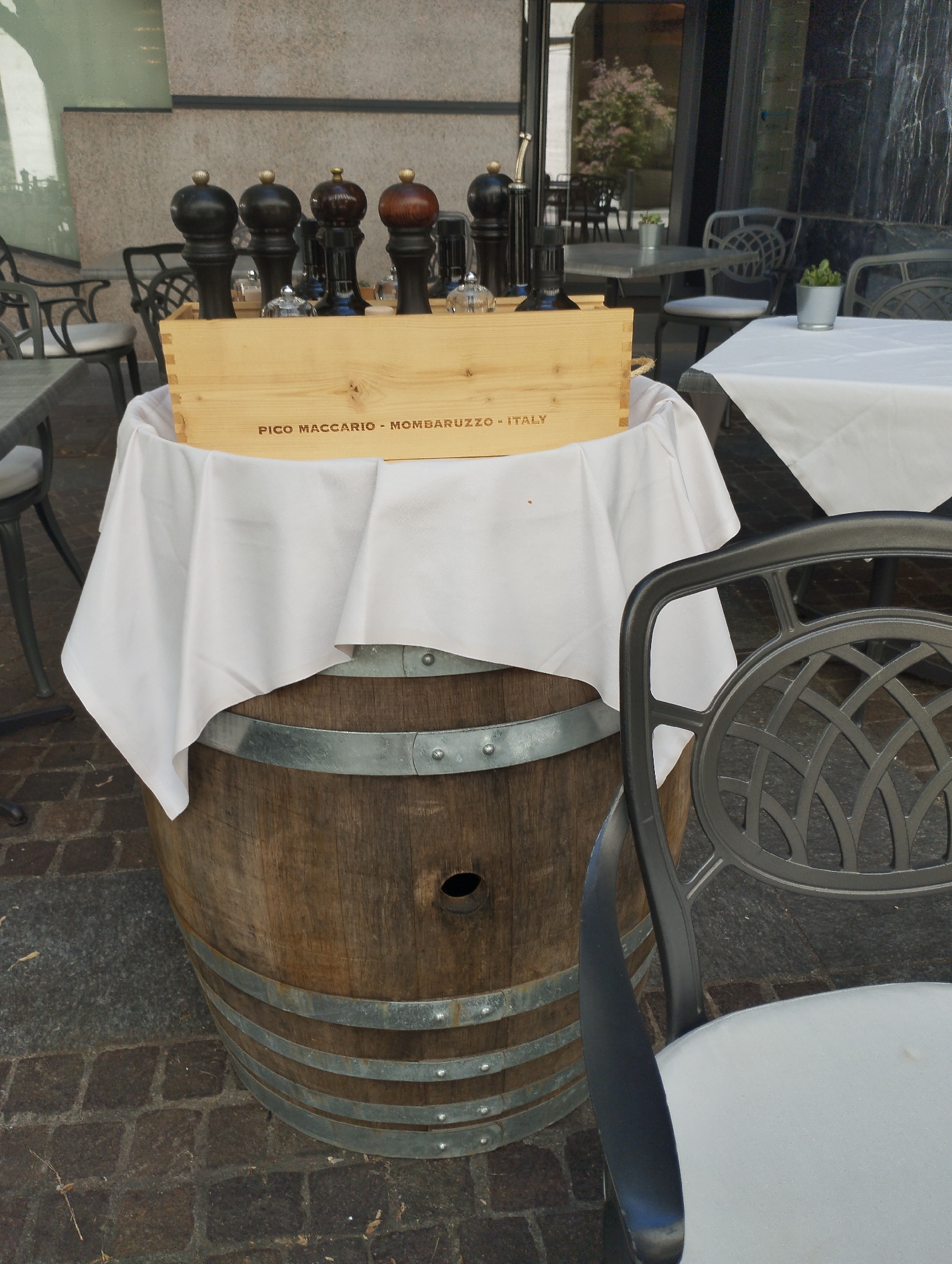 An outdoor dining setup features a wooden wine barrel as a table base, covered with a white tablecloth draped over a wooden crate. The crate displays the text "PICO MACCARIO - MOMBARUZZO - ITALY". Six large pepper grinders, some dark and some reddish-brown, are arranged in a row on top of the crate. A decorative metal chair with a white cushion is positioned to the right of the barrel. In the background, tables with white tablecloths and other metal chairs are visible, along with the facade of a building with large windows and some greenery. The scene is set on a cobblestone surface. This appears to be an outdoor seating area at a restaurant or cafe in Lugano, Switzerland, given the architectural style and the presence of outdoor dining furniture.
