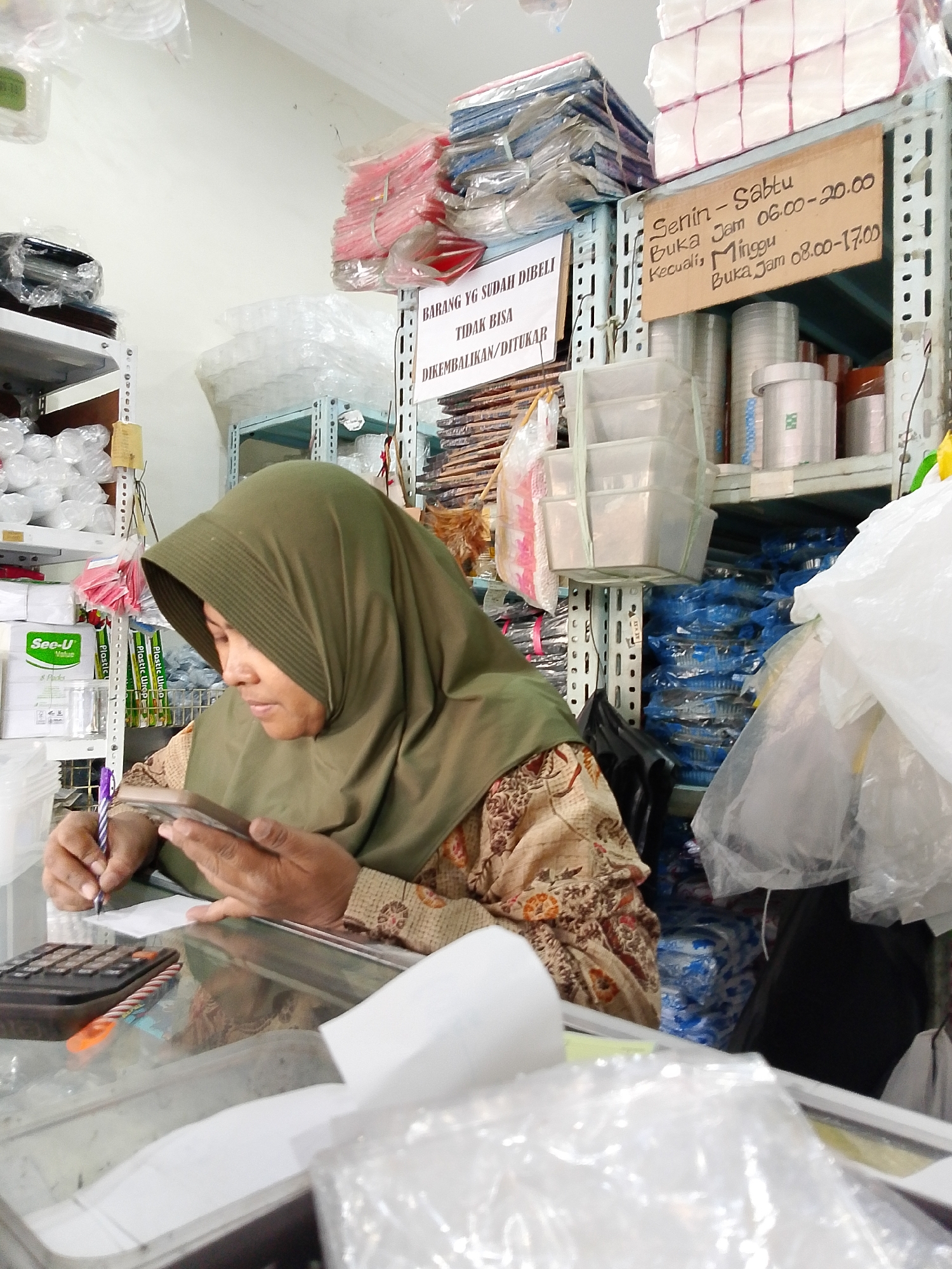 The image depicts an indoor scene, likely a small shop or storeroom, filled with various goods. The primary subject is a woman wearing a dark green hijab and a patterned garment, seated and focused on a mobile phone in her hands. She appears to be writing on a small piece of paper with a purple pen, with a calculator to her left.
The environment is characterized by shelves stacked with merchandise. On the right side of the image, a sign is visible on a shelf with handwritten text indicating business hours: "Senin - Sabtu Buka Jam 06.00 - 20.00 Kecuali Minggu Buka Jam 08.00 - 17.00". This suggests the location is a business establishment operating during specific hours, including weekends. The shelves are filled with items packaged in plastic, some appearing to be disposable containers, plastic wrap, and stacks of paper products. There are also some items in red and blue packaging. The lighting appears to be artificial, typical of an indoor retail or storage space. The overall impression is of a busy, somewhat cluttered commercial setting. Based on the signage and the context, the location is identified as Surabaya, Indonesia.