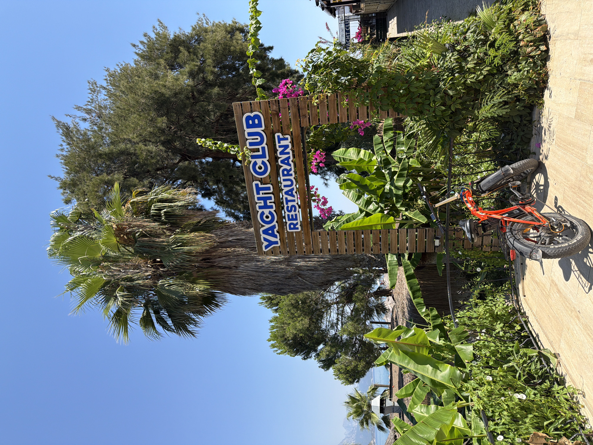 A low-angle, medium outdoor shot captures a wooden sign for "YACHT CLUB RESTAURANT" with blue lettering, positioned amidst lush tropical foliage and towering palm trees under a clear blue sky. The sign is framed by green banana leaves and vibrant pink bougainvillea flowers. A red electric bicycle with thick tires is parked in front of a black metal railing, casting a shadow on the tiled pathway. In the background, glimpses of a resort-like setting are visible, including thatched roofs and a distant view of the sea and mountains. The scene is bathed in bright sunlight, suggesting a warm, daytime environment.