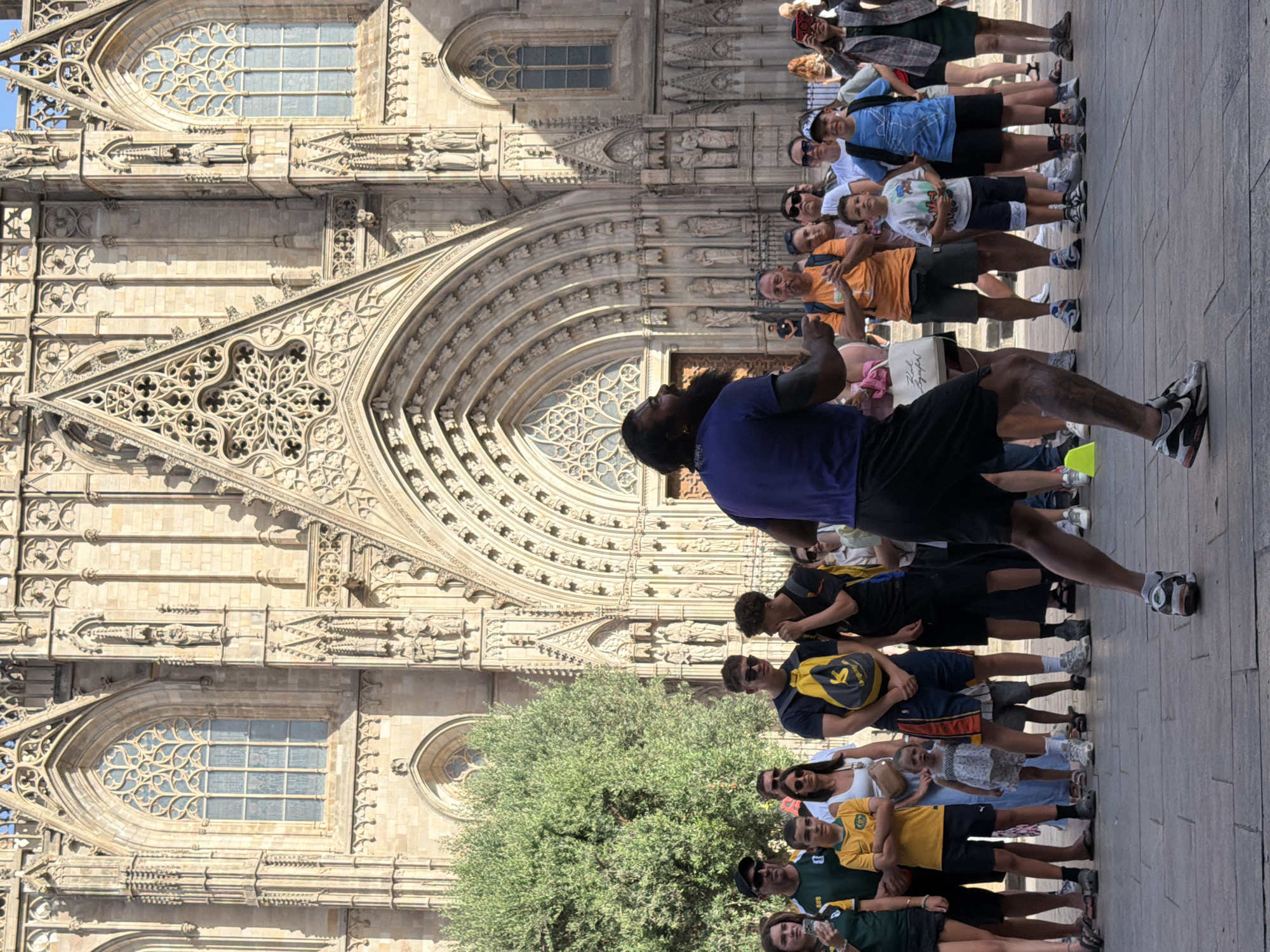 The image shows a group of people standing in a line outside a large, ornate stone building. The building appears to be a cathedral or similar religious structure, with intricate carvings, arched doorways, and stained glass windows. The architecture suggests a Gothic style, and given the context, it is likely the Barcelona Cathedral.
The people are of various ages and genders, dressed in casual summer clothing. Some are looking at the camera, while others are looking towards the building or in other directions. One person at the front of the line, wearing a dark purple shirt, is gesturing with their hands, possibly addressing the group or explaining something about the building. Several individuals are wearing backpacks or carrying bags. The time of day appears to be daytime, with strong sunlight casting shadows, indicating clear weather. The overall mood seems relaxed and tourist-like, with people enjoying the sight of the historical architecture. The image is oriented vertically, making it a portrait-style photograph.