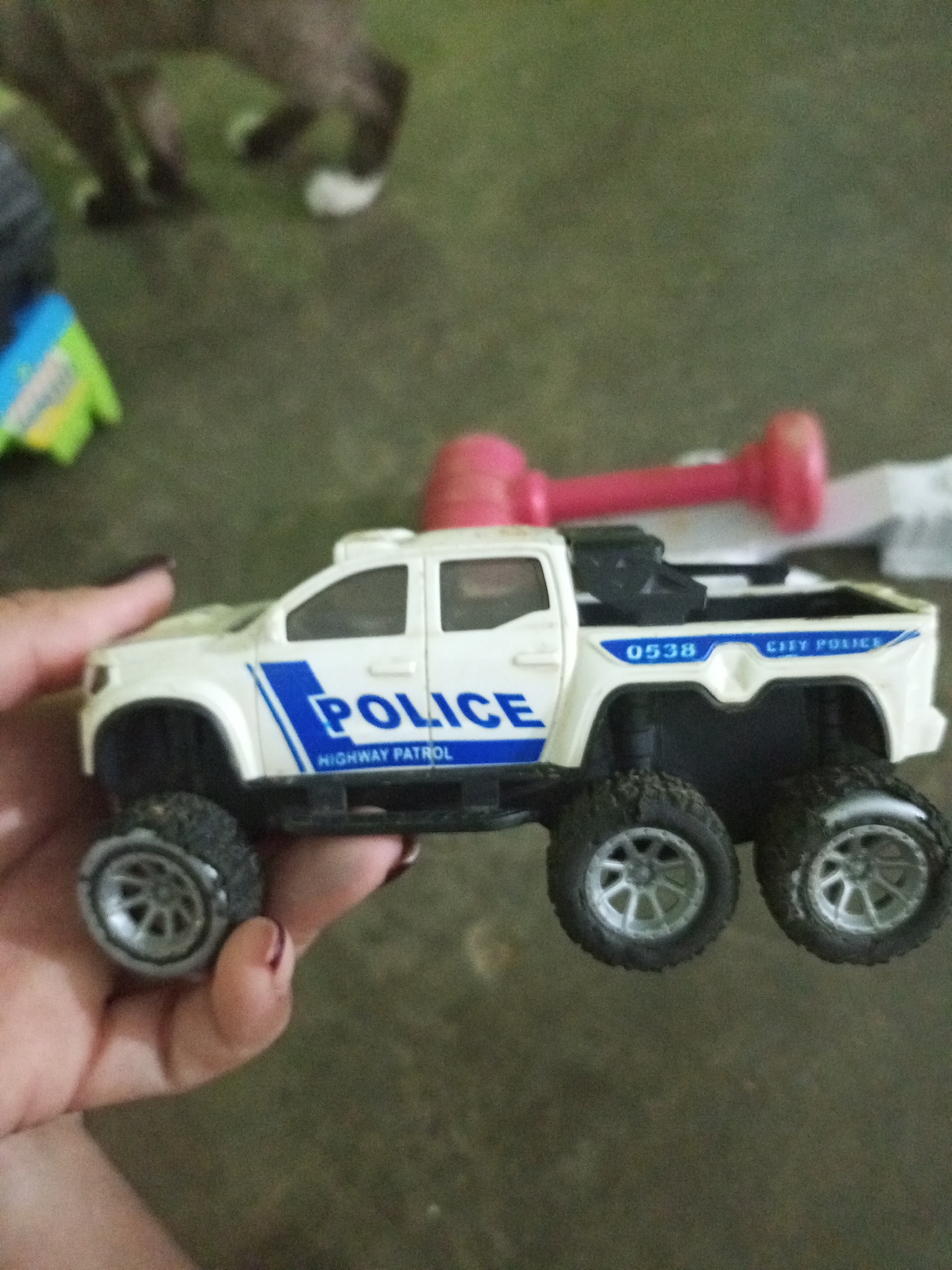 A toy police truck with "POLICE" and "HIGHWAY PATROL" written on the side is being held by a person's hand. The truck is white with blue accents and has six large, rugged tires. In the background, a blurry image of an animal, possibly a dog, can be seen. There is also a pink object resembling a toy dumbbell in the background. The ground appears to be a dirt or grassy surface. The image was taken in La Tagua, Venezuela.