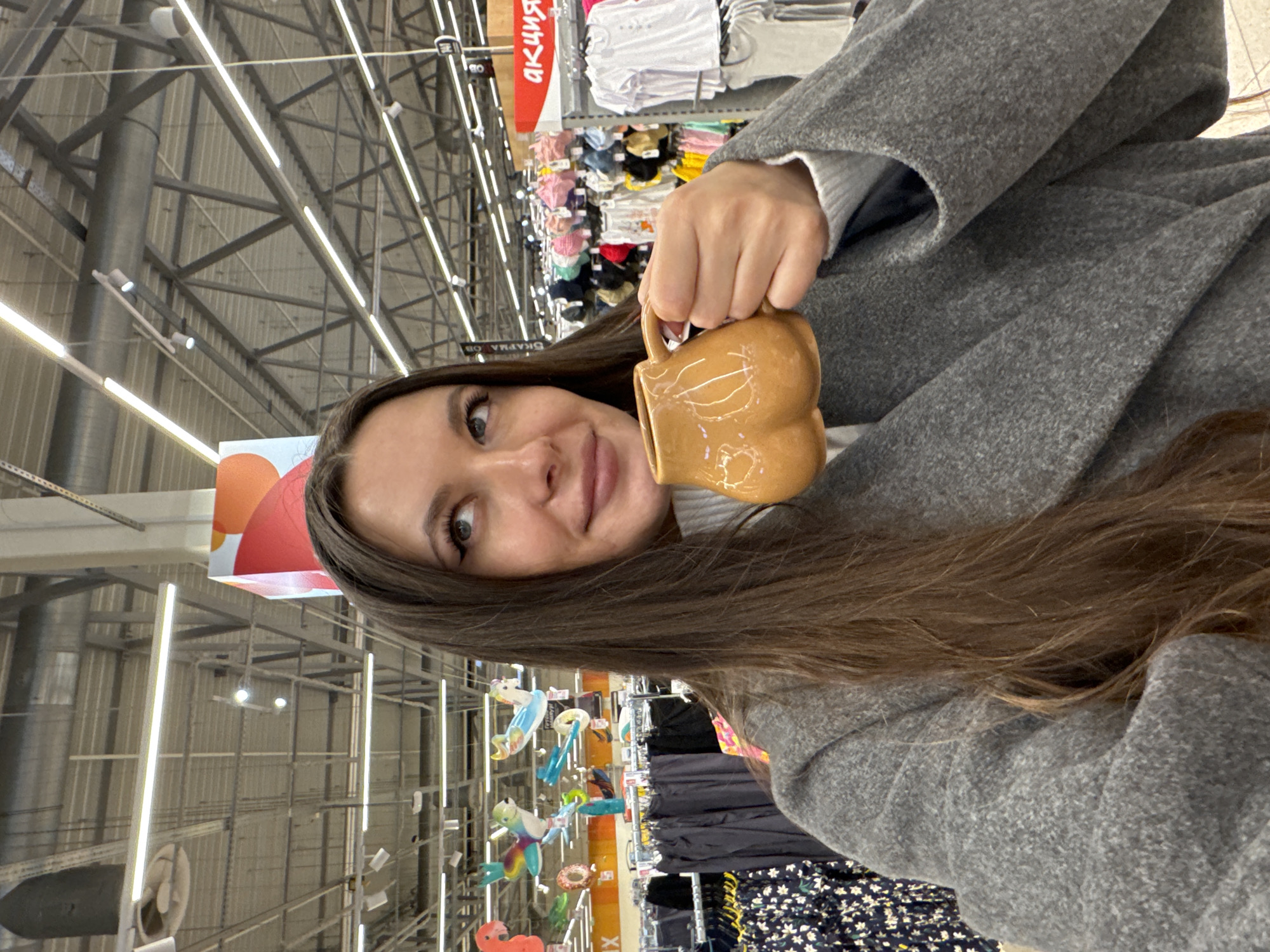 A young woman is captured in a casual portrait, likely a selfie, holding a distinctive mug in what appears to be a large retail store in Юдино, Russia. She has long, straight dark brown hair falling over her shoulders and is looking towards the camera with a subtle, pleasant smile. Her visible eye is a clear, light color, possibly green or hazel.
She is dressed in a textured grey coat or jacket, worn over a lighter-colored, possibly white, top. In her right hand, she holds a light brown, possibly ceramic or stoneware, mug. The mug features a unique, raised, sculpted design on its side, which appears to be a stylized heart or organic shape, and has an ear-shaped handle.
The background indicates a spacious commercial environment, likely a supermarket or hypermarket. Rows of shelving stocked with various merchandise, including colorful children's clothing or baby items, are visible. The ceiling exhibits an industrial aesthetic with exposed silver-toned beams, large ventilation ducts, and numerous bright linear LED light fixtures. Hanging above the aisles are colorful inflatable decorations, such as a unicorn, a donut, and a flamingo, suggesting a section catering to children's goods or summer items.
Visible text on red signs in the background includes the Cyrillic words "АКЦИЯ" (AKCIYA), meaning "Sale" or "Promotion," and "СКИДКИ" (SKIDKI), meaning "Discounts," reinforcing the retail setting. The overall mood of the image is relaxed and candid.