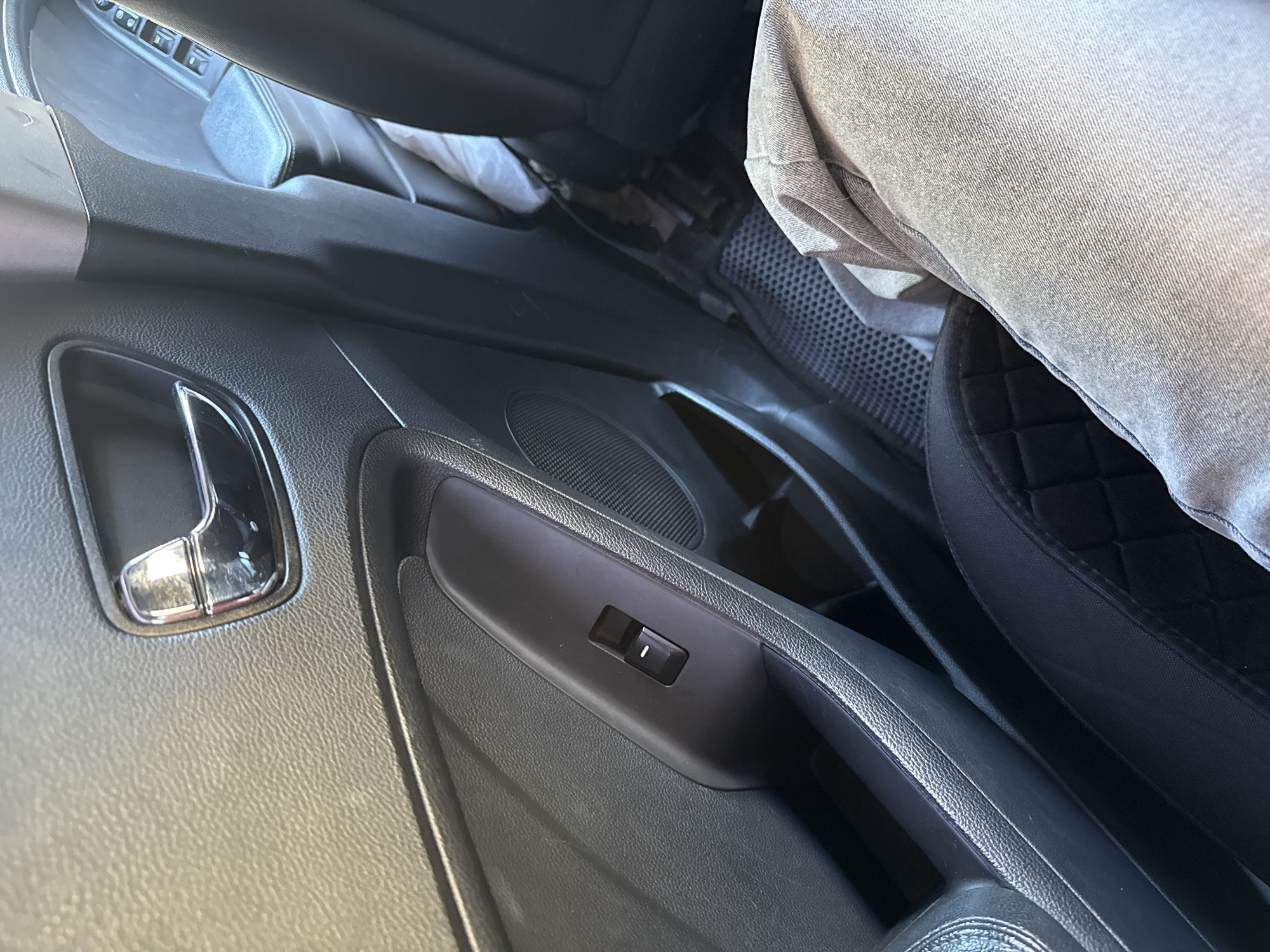 This image captures a close-up view of the interior of a car, likely in Saint Petersburg, Russia. The primary subjects are parts of the car's interior and a person's leg. We see a textured black door panel with a chrome door handle, a window control switch with a small white indicator, and a speaker grille. To the right, a person's leg, clad in light grey denim or similar fabric, rests on the car seat. The seat itself has a quilted black upholstery visible. The scene is well-lit, suggesting daytime, with natural light entering the car. The overall impression is one of a mundane, everyday moment inside a vehicle, possibly during transit or while parked. There is no visible text within the image. The focus is on the tactile textures of the car's interior materials and the casual posture of the person.