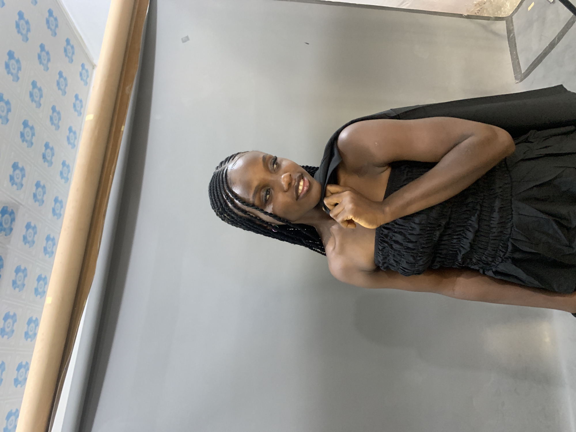 A young woman with braided hair is posing for a photo shoot in front of a gray backdrop. She is wearing a black strapless dress with a ruched bodice and a gathered skirt. She has a black jacket draped over her left shoulder. Her head is turned to her right, and she is looking slightly down and to the side with a subtle smile. The background appears to be indoors, with a rolled-up brown paper backdrop at the top and a section of a gray paper backdrop visible. The ceiling has a tiled pattern with blue floral designs. The floor is not clearly visible but appears to be a studio floor. The lighting suggests an indoor studio setting, possibly in Gura Top, Nigeria.