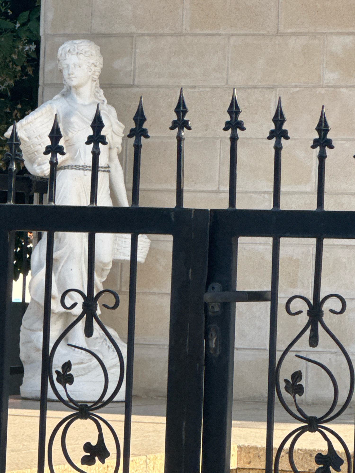 The image shows a partial view of a white marble statue of a woman holding a basket, visible behind a black wrought-iron gate. The statue is located in front of a light-colored brick wall. The gate has decorative elements including pointed finials and scrollwork. The lower portion of the statue shows the woman's draped clothing and the basket she is holding. The scene appears to be outdoors, with sunlight casting shadows. The location is identified as Mesa Geitonia, Cyprus.