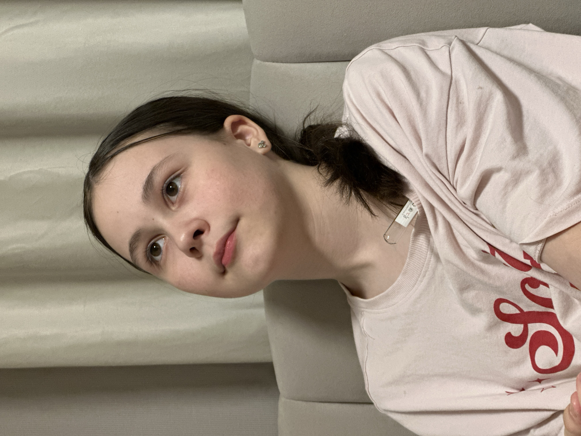 A young, white female lies on a light-grey upholstered sofa in an indoor setting. She has dark hair pulled back in a low ponytail, fair skin with slightly flushed cheeks, and light-brown eyes. Her lips are pink, and she wears a small, clover-shaped earring in her left ear. She is wearing a light-pink t-shirt with cursive, red writing. The background includes a neutral, possibly white, curtain or wall. No information is available to ascertain the location in Votkinsk, Russia.