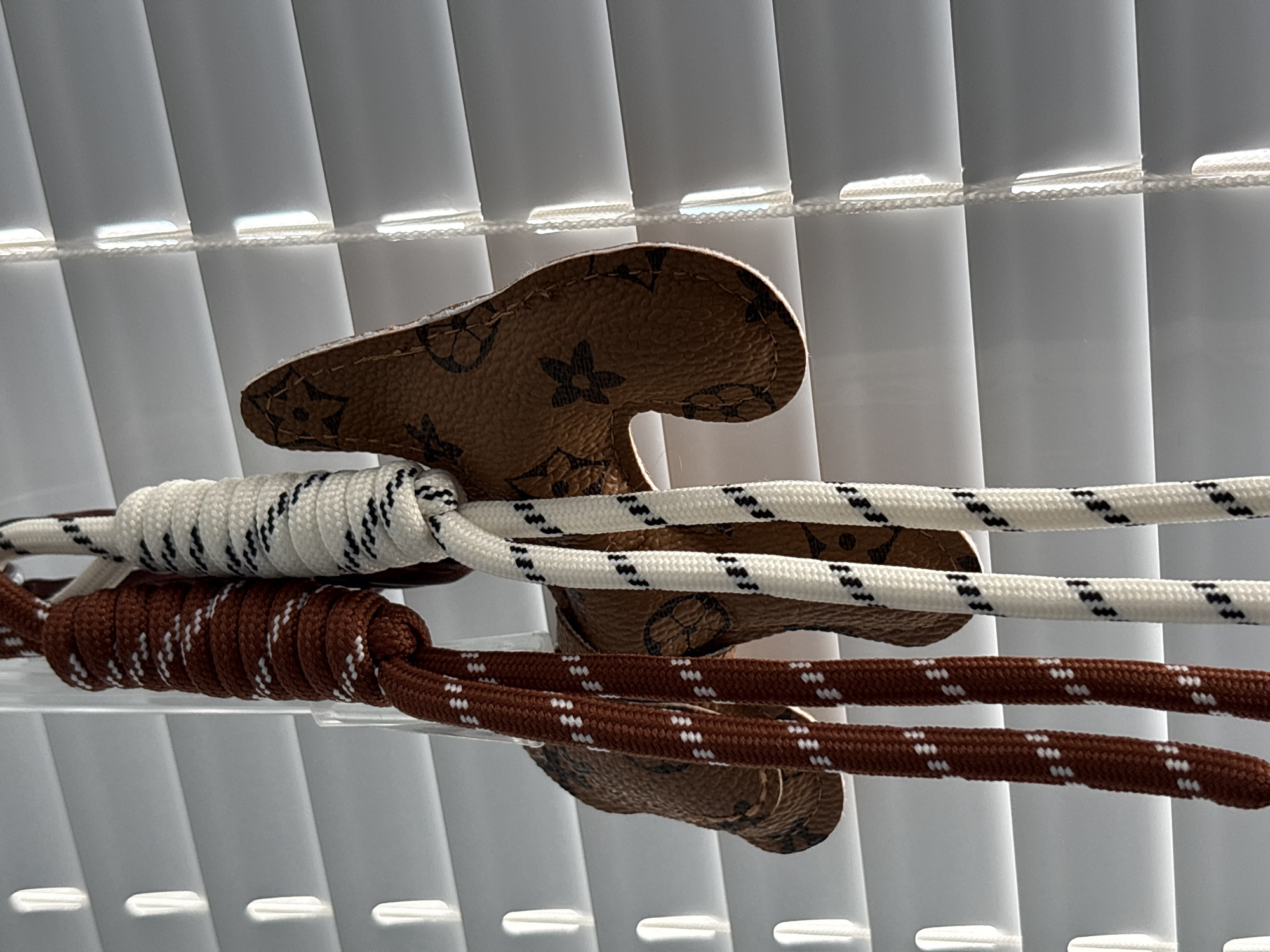 This close-up image, set in Sofia, Bulgaria, features two thick, textured ropes and a shaped, monogrammed object, all positioned horizontally against a backdrop of white vertical window blinds. The upper rope is white with distinct black zigzag stripes, while the lower rope is brown with a pattern of white dashed lines or dots. Both ropes exhibit a section where they are tightly coiled or wrapped around themselves or an unseen core, creating a textured, decorative effect.
Behind these ropes is a light brown, leather-like material, possibly synthetic, which displays a repeating pattern strongly resembling the iconic Louis Vuitton monogram, including stylized floral elements and the "LV" initials. This object has a distinct, somewhat organic shape, possibly a decorative tab or part of a larger item, and shows visible stitching along its perimeter.
The background consists of modern, white vertical blinds, which are partially open, allowing bright daylight to enter and create prominent horizontal lines of light where the individual slats are connected by fine cords. The lighting indicates it is daytime. The scene appears static and decorative, with no human presence or overt activity.