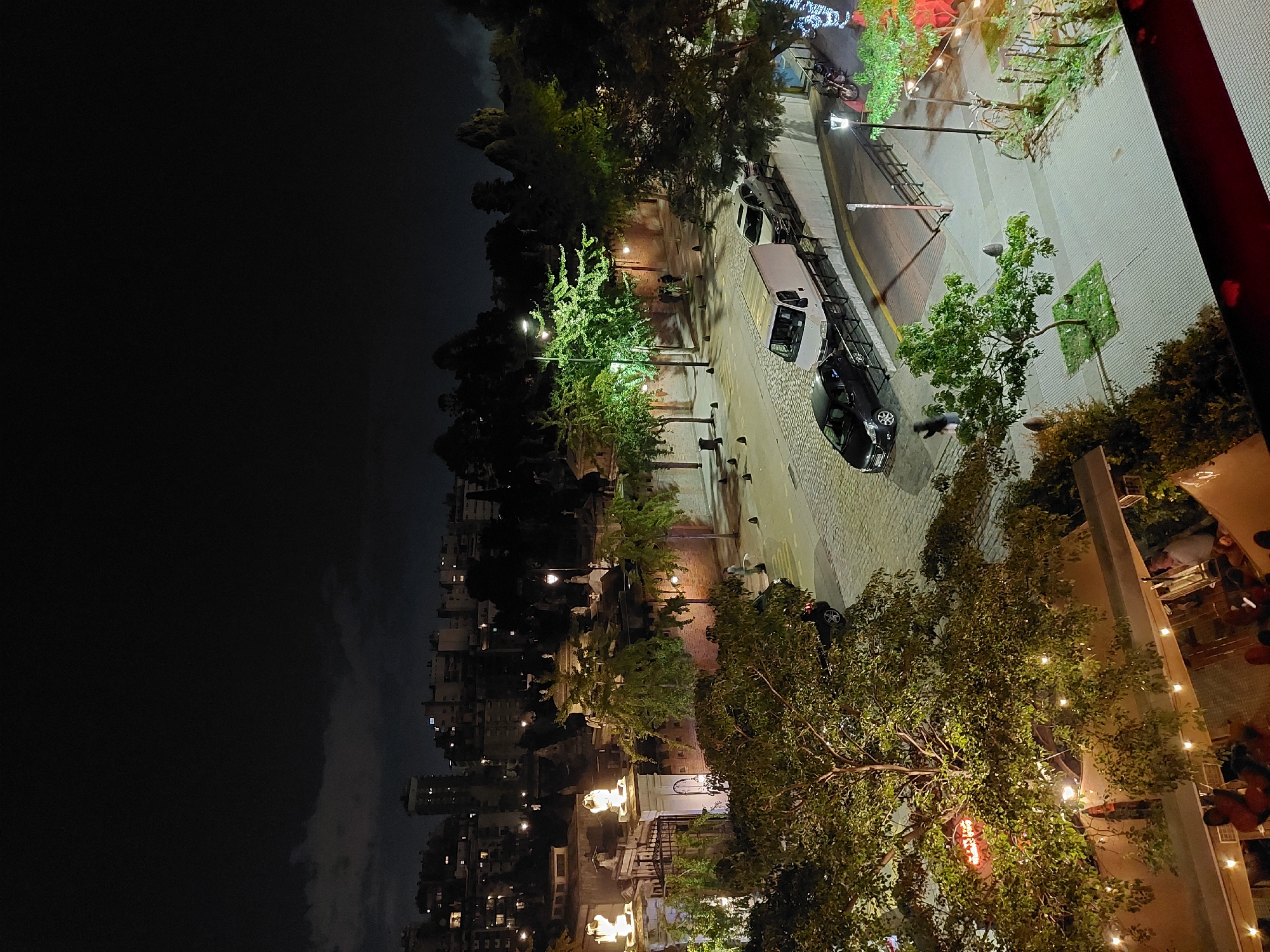 The image presents a night scene in a lively urban environment, likely Buenos Aires, Argentina. It's an elevated perspective, possibly from a balcony, overlooking a street with pedestrian activity and vehicular traffic.
People are visible in small groups, seemingly in outdoor dining areas. There are vehicles parked along the street, including a white van. Buildings line the street, extending into the distance where they are illuminated by streetlights and building lights. There are trees adorned with string lights, adding to the ambiance. To the left, there is an area of relative darkness, possibly a park or open space, with distant city lights visible against the night sky. The street surface appears to be paved, and the overall scene conveys a bustling urban atmosphere.