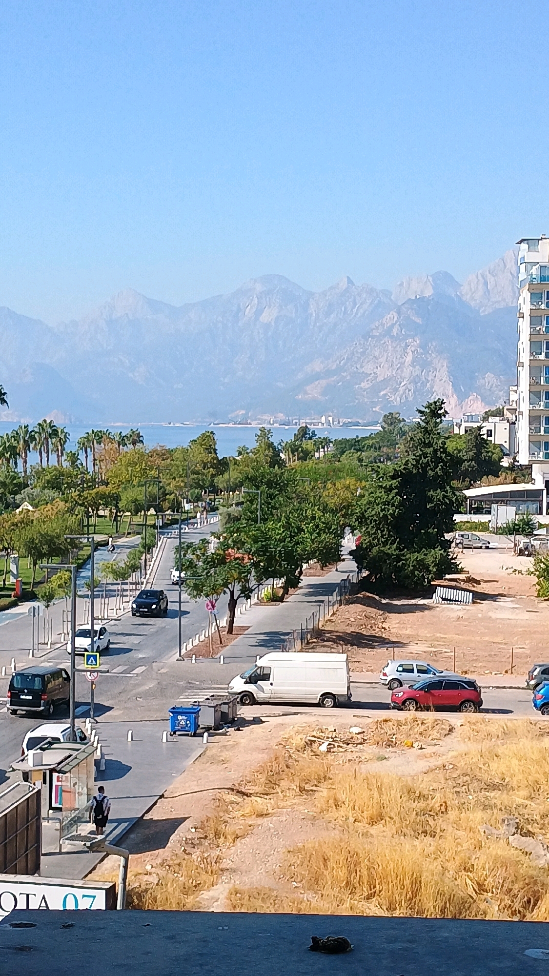 The image captures a sunlit urban scene in Konyaaltı, Turkey, characterized by a wide, paved road flanked by trees and sidewalks. In the foreground, a portion of a building's roof is visible, with the partial text "OTA 07" on its side. Across the street, a bus stop with a person waiting is located. A black van is parked nearby, and a white car is also present. Further down the road, a white van drives, followed by a red SUV and a blue car. A blue recycling bin and two other containers are situated on the sidewalk. The road leads towards a distant, hazy coastline with palm trees and a large body of water. In the background, a mountain range stretches across the horizon, with a modern, multi-story building visible on the right side of the frame. The sky is clear and blue.