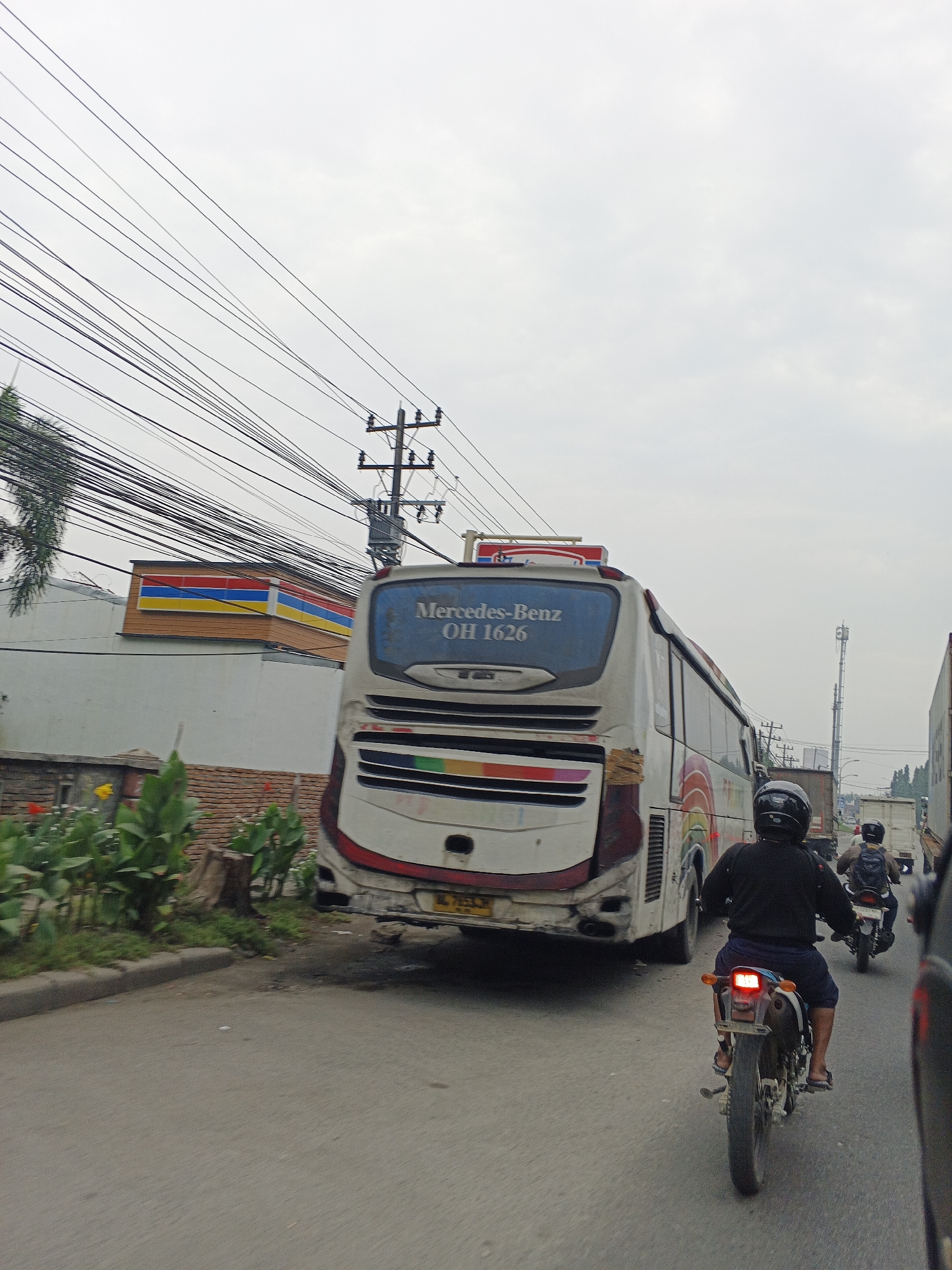 The image depicts a street scene in Tg Morawa A, Indonesia, focusing on the rear of a white bus with "Mercedes-Benz OH 1626" written on its back window. The bus has a rainbow-colored stripe along its side and is moving on a paved road. In front of the bus, a motorcyclist wearing a black helmet and dark clothing rides a dirt bike with a glowing red taillight. Behind the bus, another motorcyclist with a backpack is visible. The road is flanked by a sidewalk and some greenery, including plants and a tree stump. In the background, there is a building with a colorful facade resembling a convenience store, overhead power lines, utility poles, and other vehicles, including a truck. The sky is overcast.