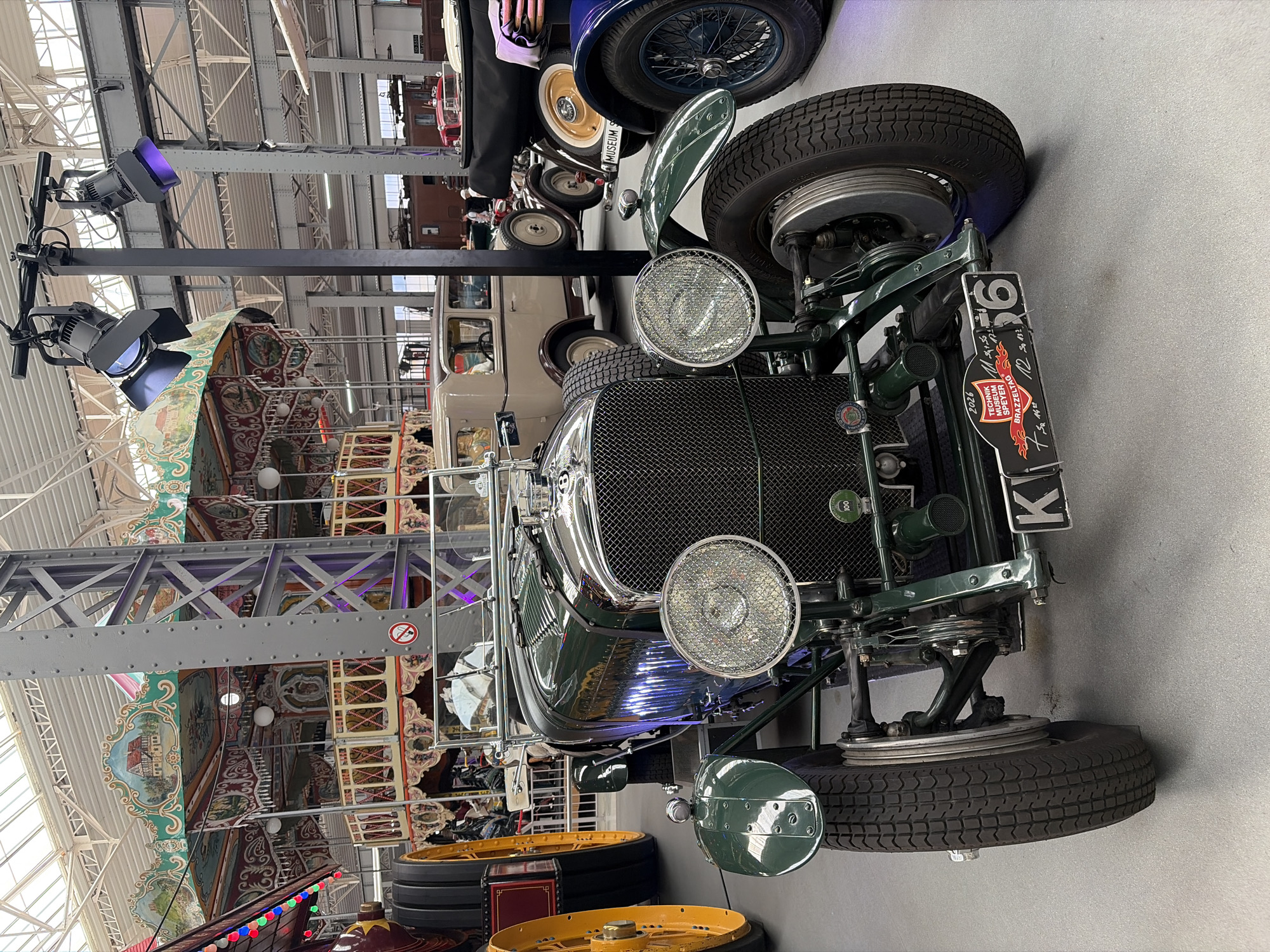 The image shows a vintage green car parked indoors in the Technik Museum Speyer. The car is the primary subject, positioned in the foreground and taking up a significant portion of the frame. It features a prominent chrome grille, large circular headlights with wire mesh covers, and visible suspension components. A license plate reading "K 56" is attached to the front bumper, along with event stickers, one of which says "2026 TECHNIK MUSEUM SPEYER BRAZZELTAG". The car's front wheel is turned outwards.
In the background, other vehicles are visible, including a light-colored sedan and parts of other cars, some appearing to be older models as well. A carousel with painted panels and decorative elements is also visible behind the vintage car, suggesting the museum might have various themed exhibits. Overhead, industrial-style lighting fixtures are mounted on poles, casting colored light, including blue and purple, onto the scene. The ceiling appears to be a glass or translucent material supported by a metal framework. The floor is a light grey, polished surface. No people are clearly visible in the immediate vicinity of the green car, though figures might be present further in the background. The overall environment suggests an indoor exhibition space within a museum.