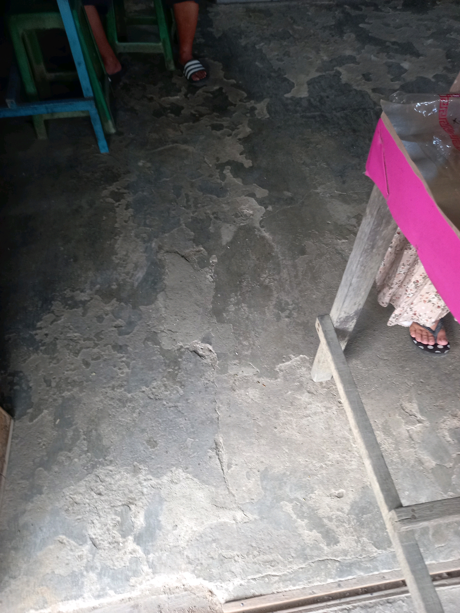 The image shows a low-angle, eye-level view of a concrete floor. In the upper left, a person's legs are visible, wearing black flip-flops with white stripes. They are sitting on a green chair with blue metal accents. To the right, another person's legs are visible, wearing a floral-patterned dress and dark flip-flops with white dots. They are seated near a wooden table with a bright pink tablecloth. The environment appears to be an outdoor or semi-outdoor space, possibly a market or a roadside eatery, given the rustic furniture and concrete flooring. The concrete itself is worn and uneven, with patches of lighter and darker coloration, suggesting exposure to the elements. There are no distinct urban features visible that would specifically place it in Samarinda, Indonesia, beyond what was provided.