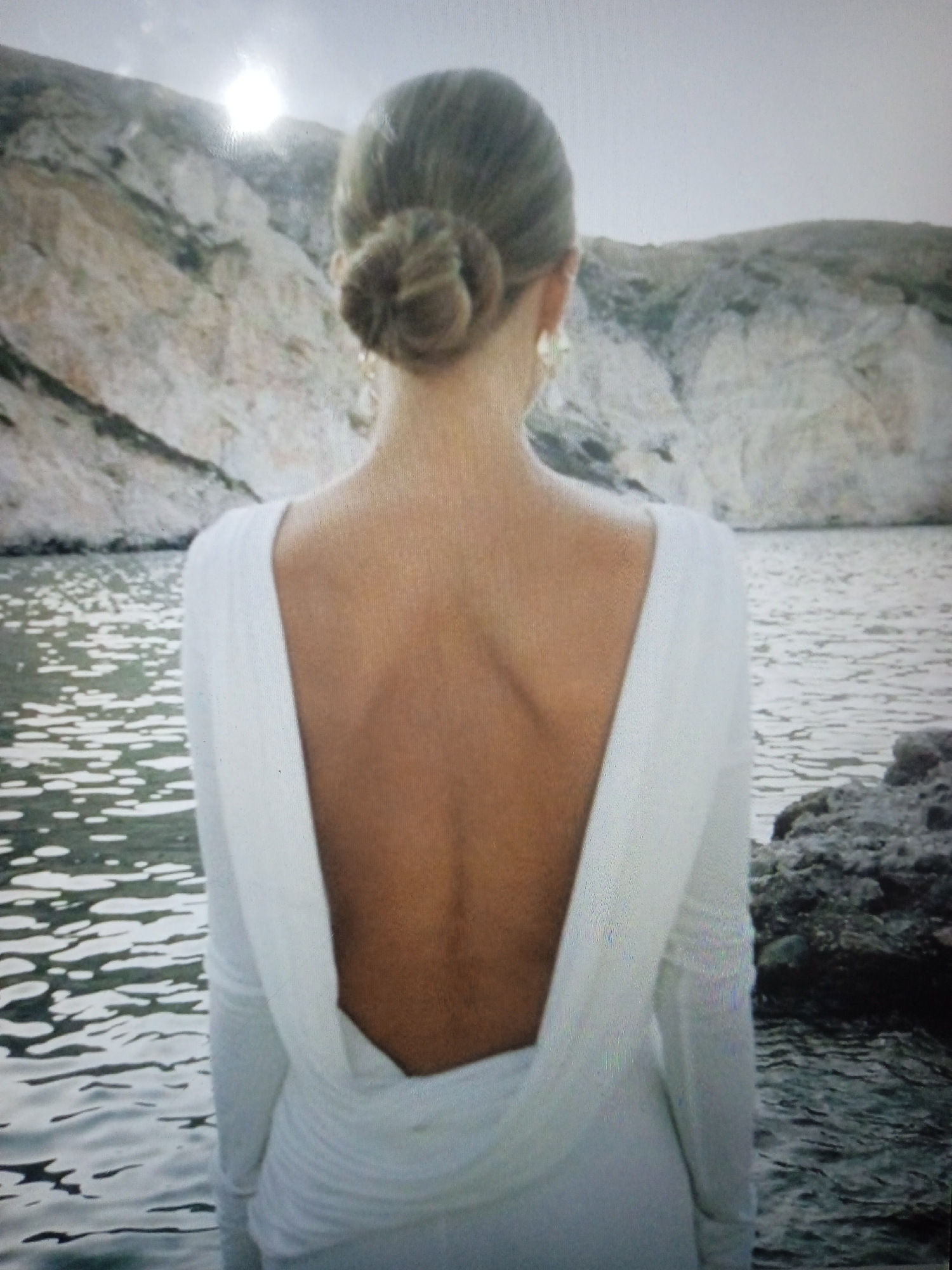 The image shows the back of a woman wearing a white, long-sleeved dress with a very low, open back. Her hair is pulled up in a bun. She is standing outdoors near a body of water, with rocky hills in the background. The sun is visible in the upper left corner. The scene is a natural landscape, not a city environment. The image does not provide any information about the city of Chernivtsi, Ukraine.